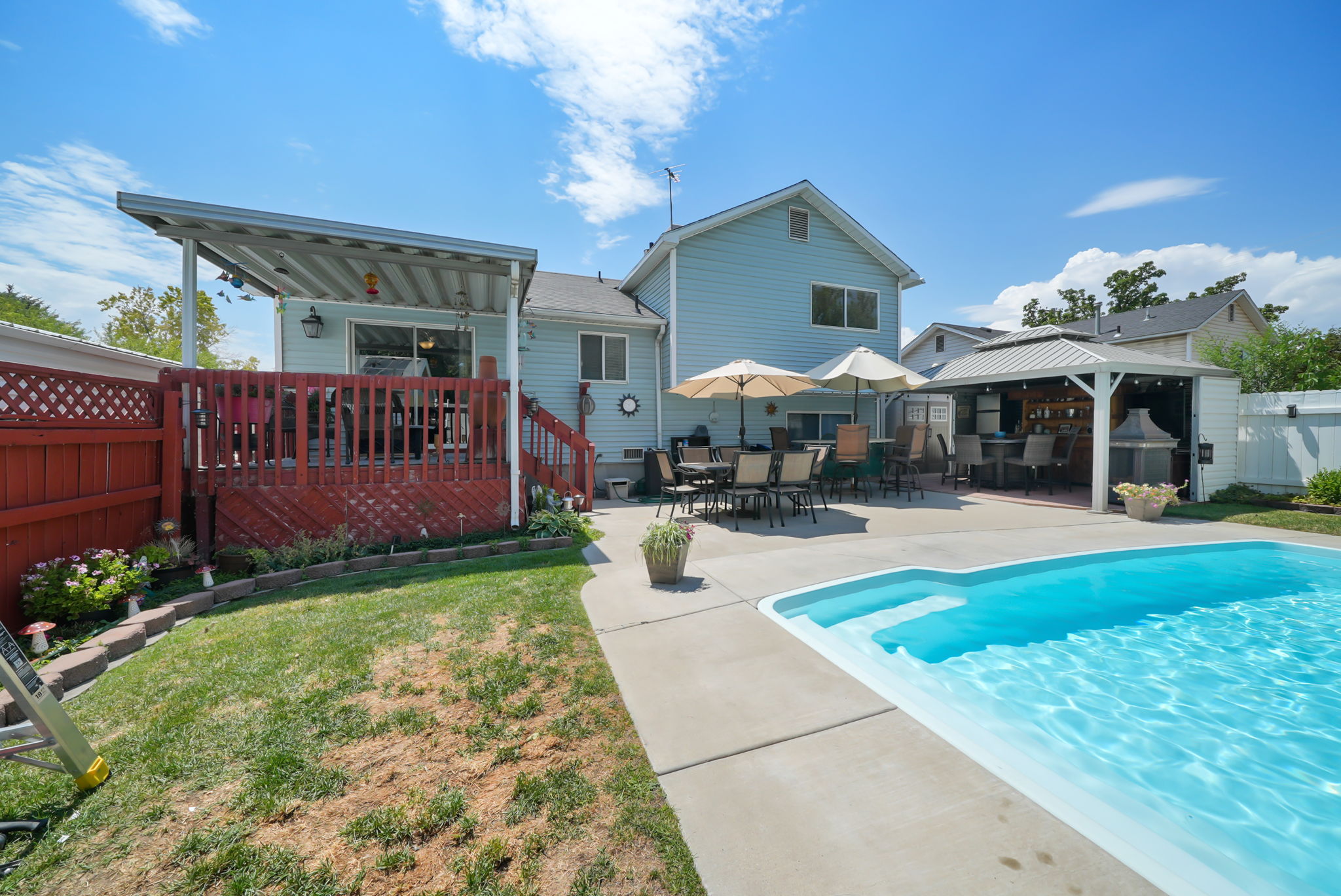 Expansive backyard featuring a fenced-in swimming pool, a covered wood-finish deck with stairs, a built-in outdoor bar with a pergola, and concrete patio areas