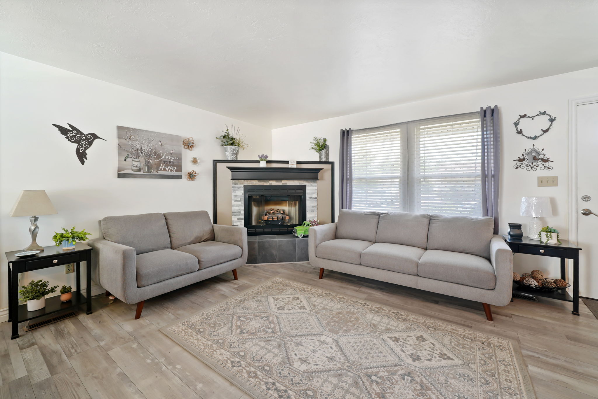 Living room featuring a central fireplace with a stone surround and dark mantel, light wood-finish flooring, and a large window with blinds