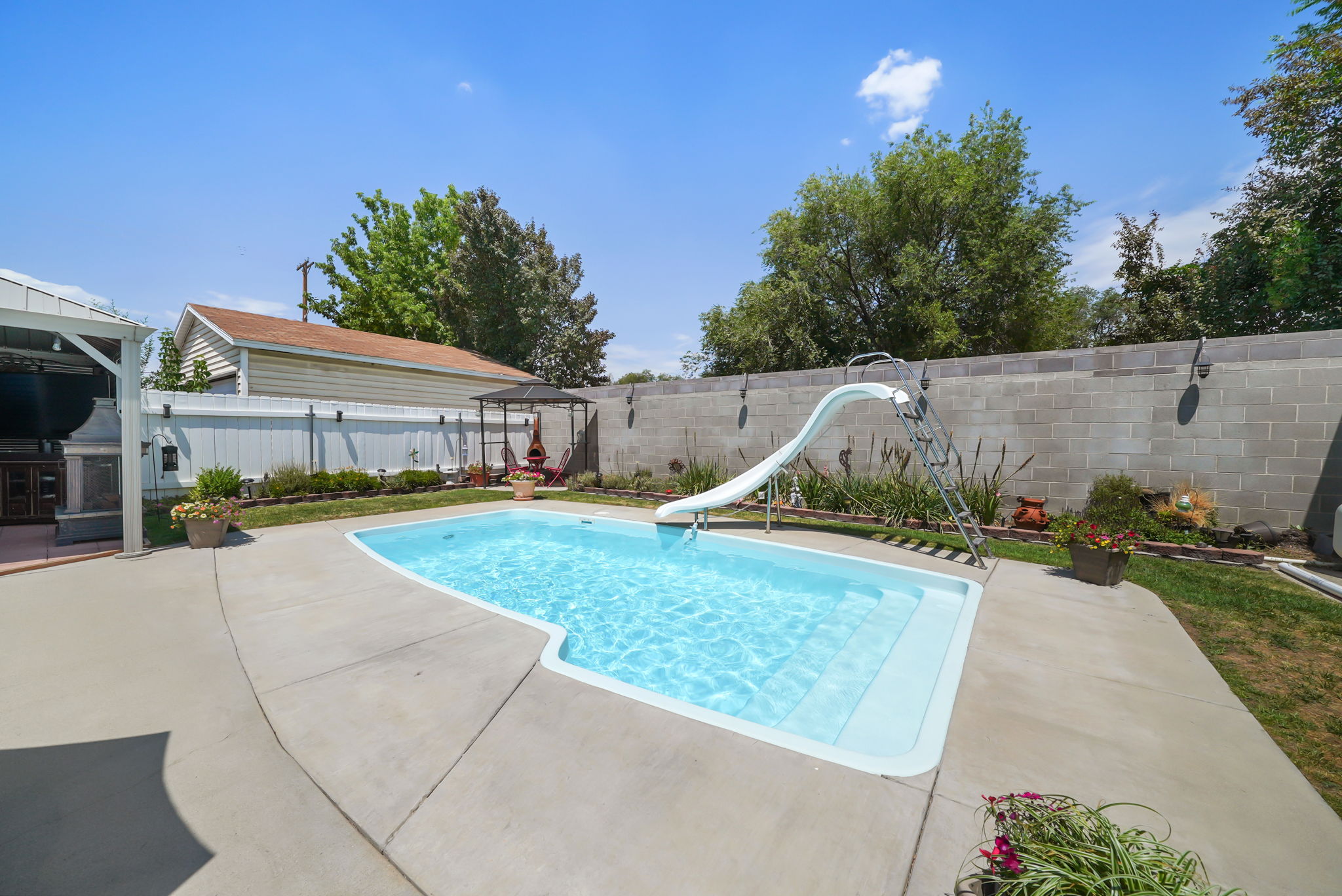 Backyard oasis featuring an in-ground swimming pool with a slide, extensive concrete patio areas, a built-in outdoor kitchen under a pergola, and a private perimeter fence
