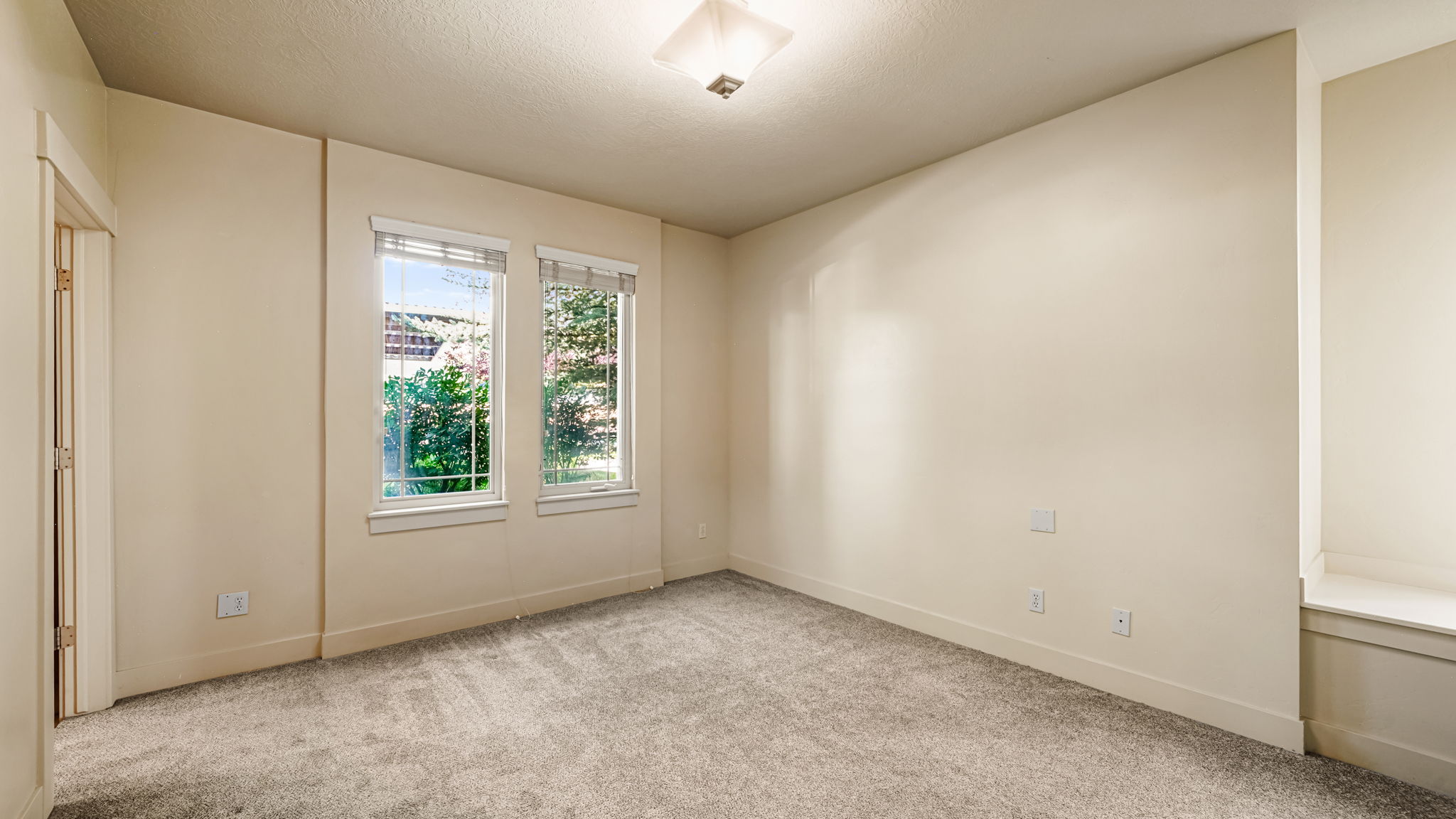 Neutral-toned room featuring light gray carpet, dual windows with blinds, a built-in window bench, and a ceiling-mounted light fixture