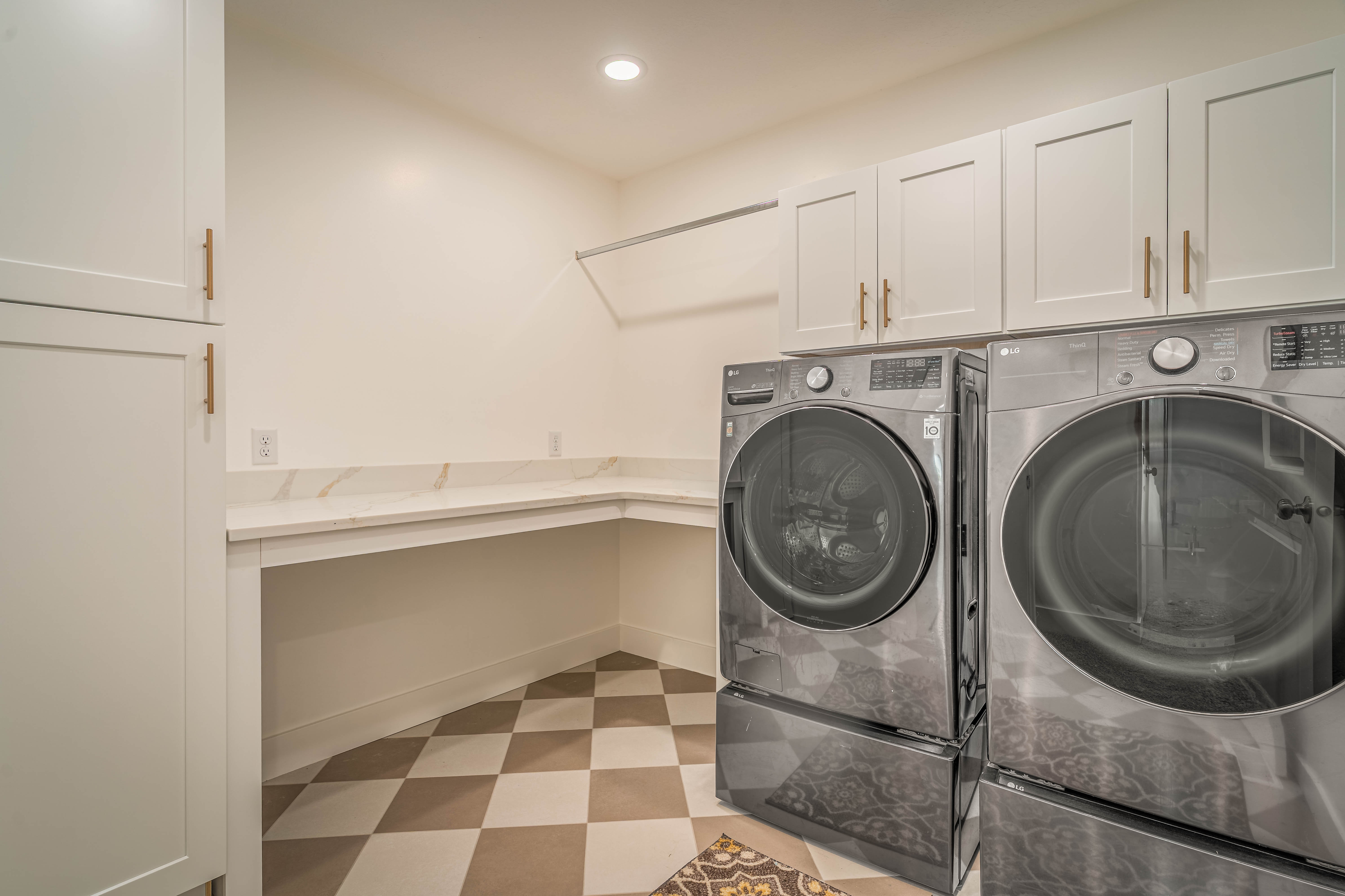 Laundry room featuring a white countertop with a partial backsplash, white shaker-style cabinetry with brass hardware, and checkered floor tiling