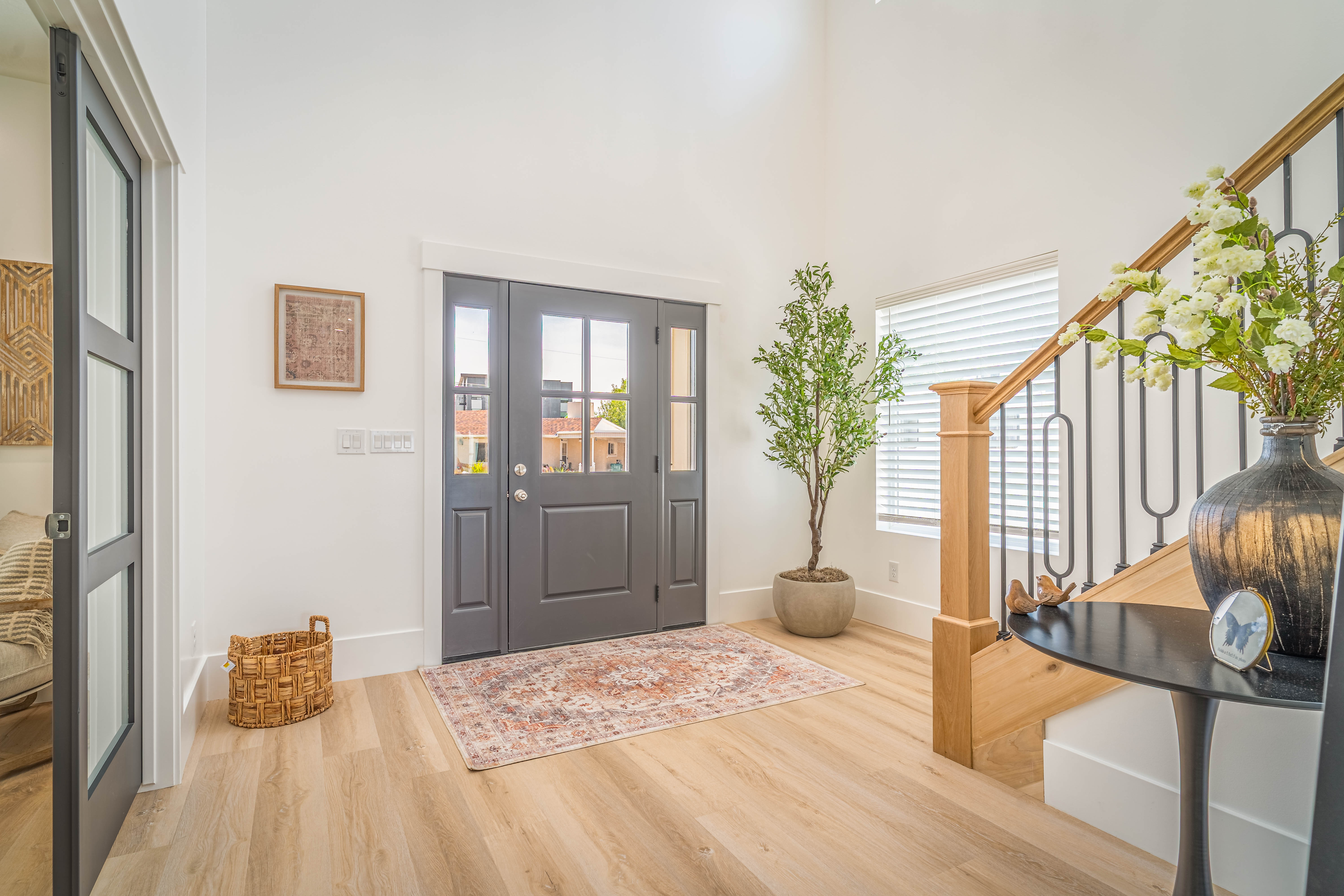 Entryway featuring light wood-finish flooring, a contemporary dark gray front door with glass panels, a white accent wall, a staircase with a natural wood handrail and black iron balusters, and a side window with horizontal blinds