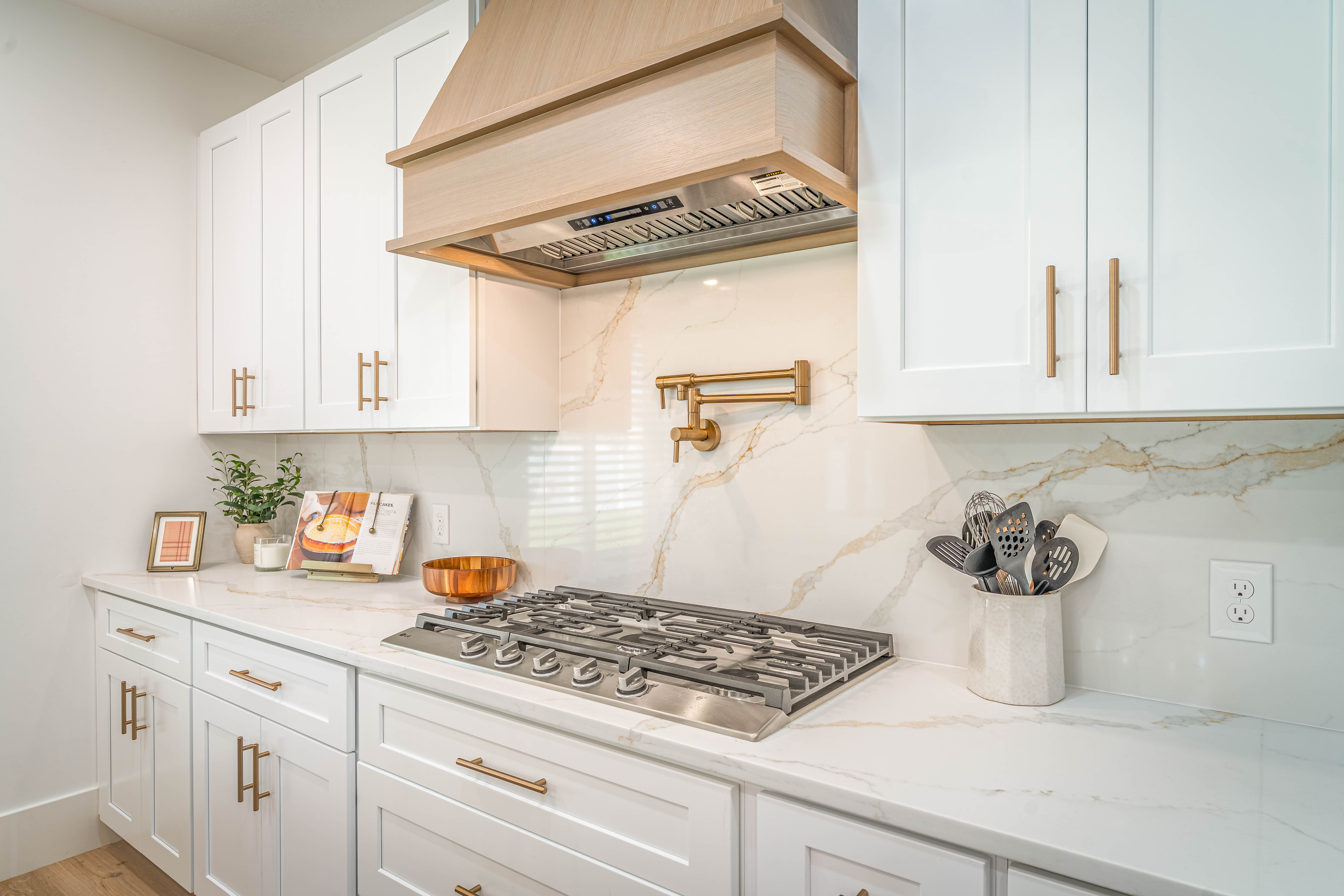 Kitchen featuring white shaker cabinetry, white countertops with veining, a stainless steel gas range, a wood-finish range hood, and a brass-finish pot filler