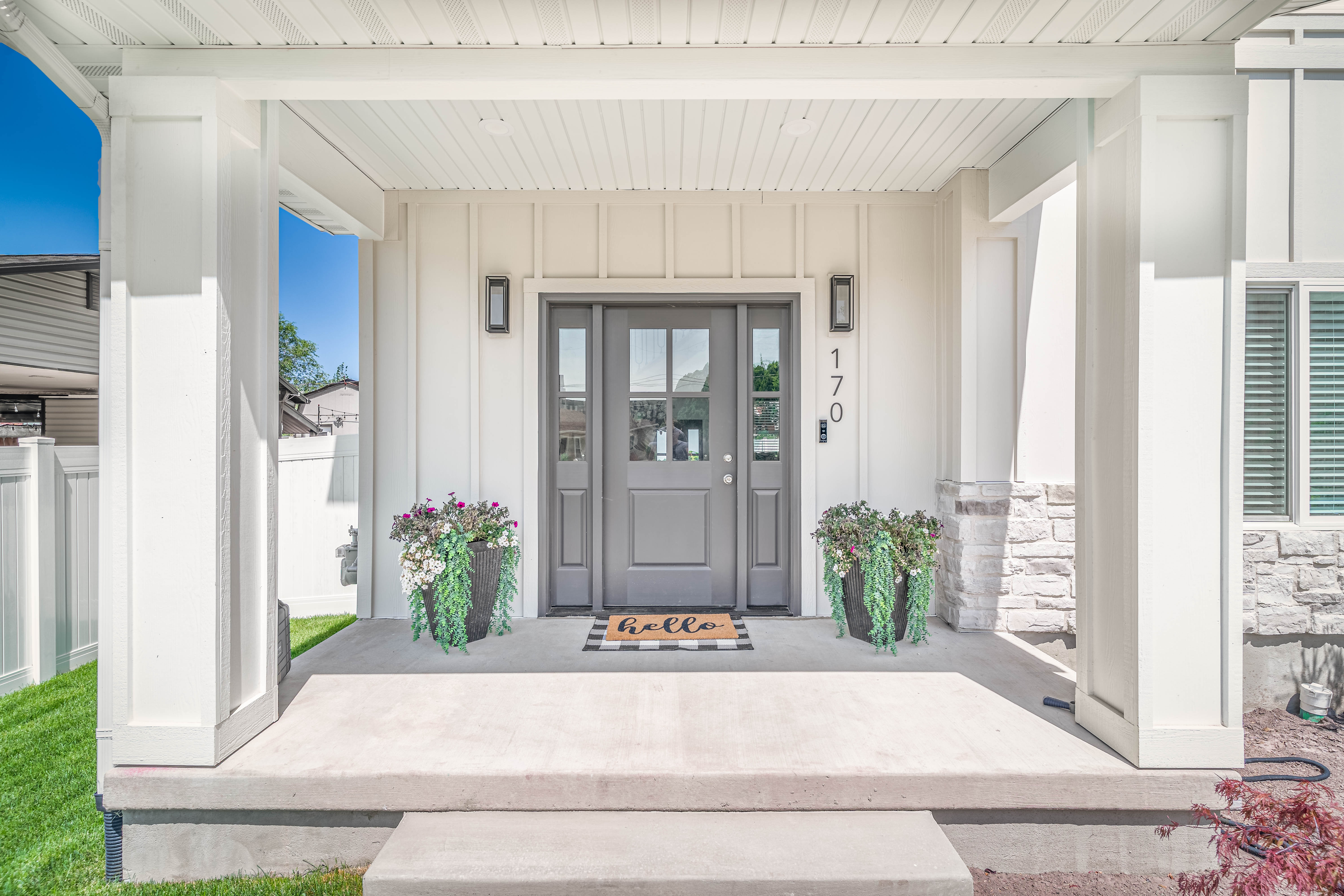 Covered front porch featuring a gray entry door with glass panels, vertical siding, stone accents, and two modern outdoor sconces