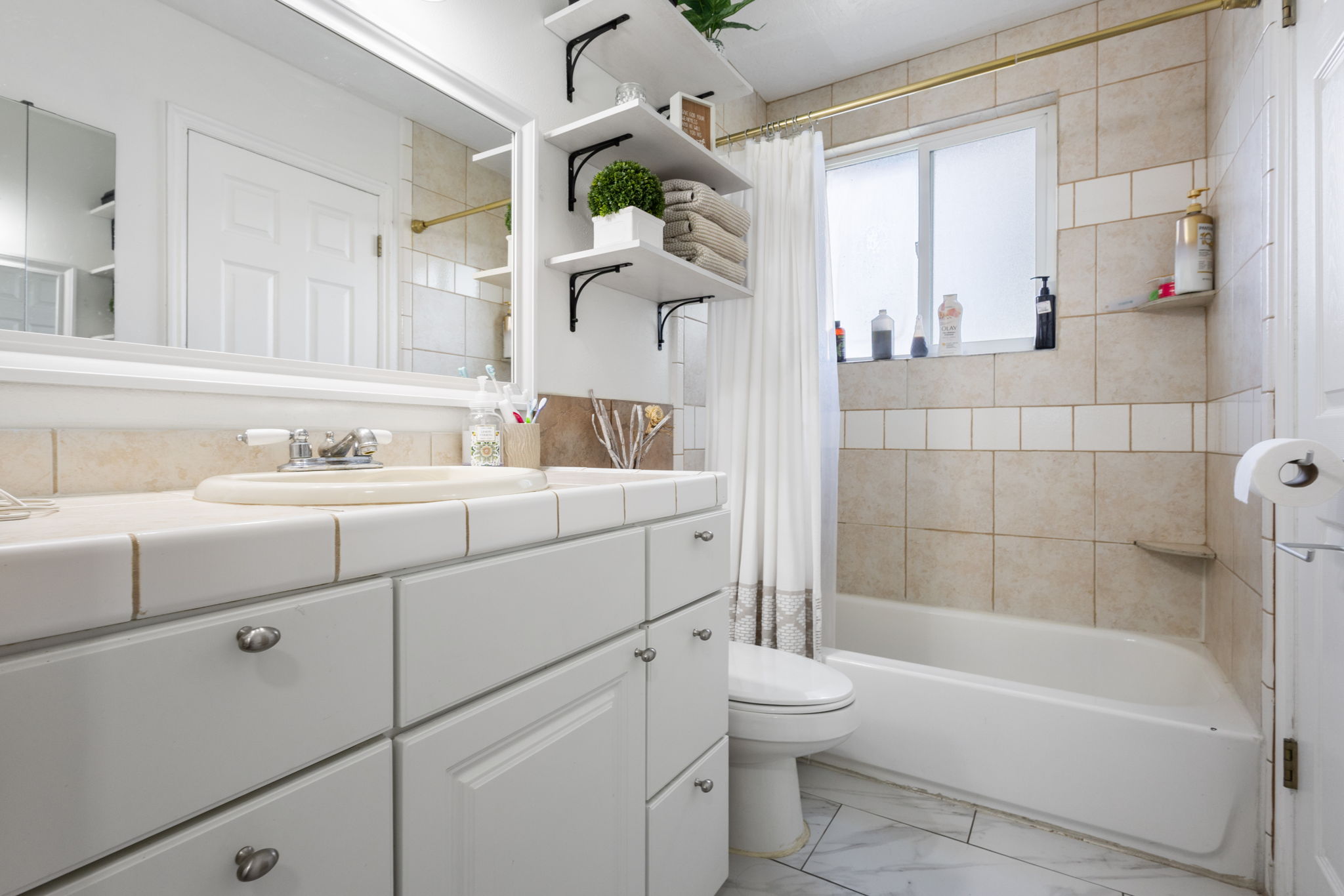 Vanity with integrated sink and tile countertop, white cabinetry with silver hardware, large framed mirror, built-in shelving, tub with tiled surround, and a frosted window