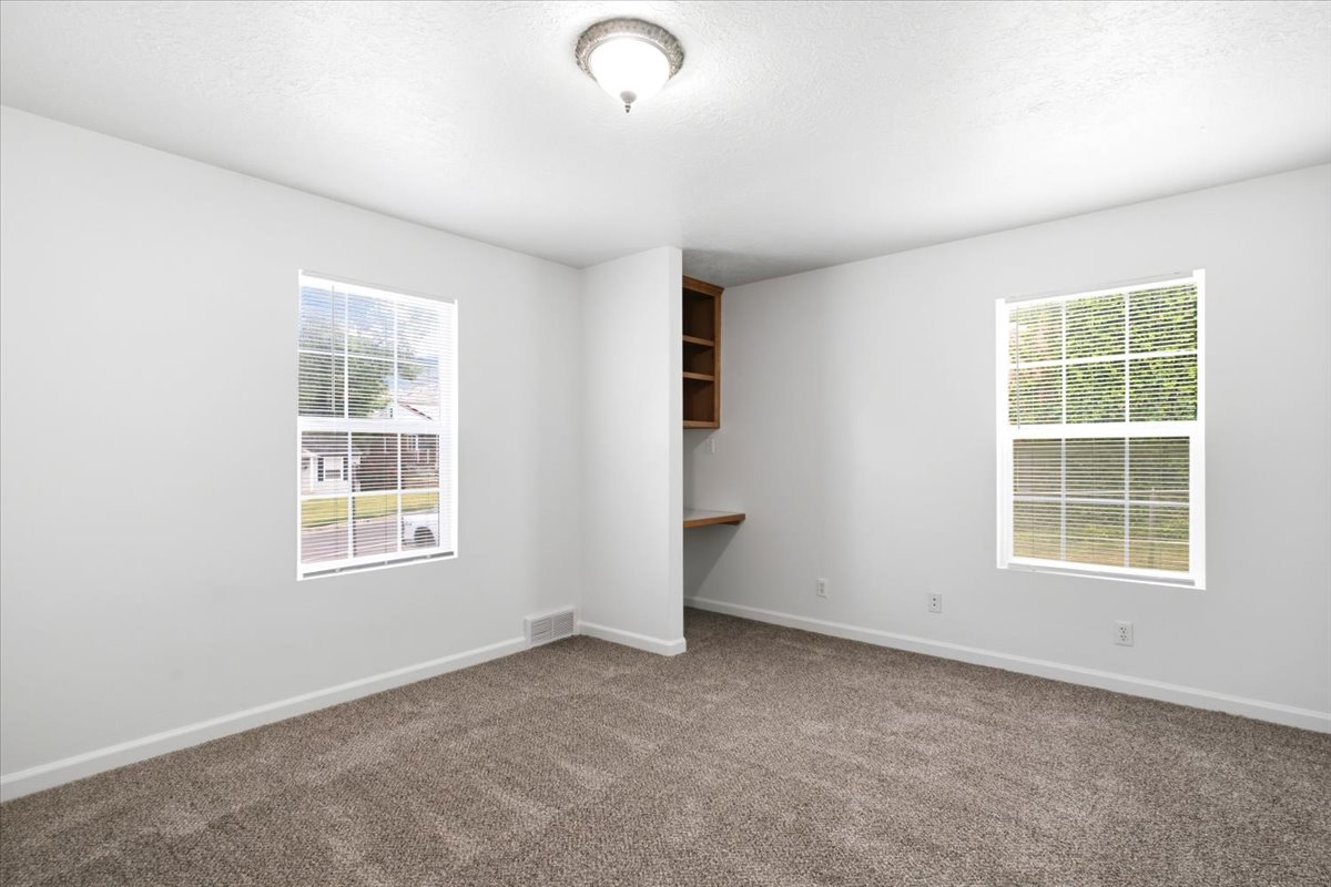 Bright interior room with plush carpeting, two windows, and a built-in wooden desk with shelving