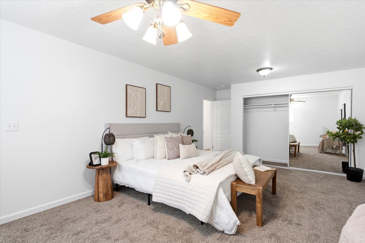 Carpeted bedroom featuring a light gray upholstered headboard, white walls, and a ceiling fan with light fixture