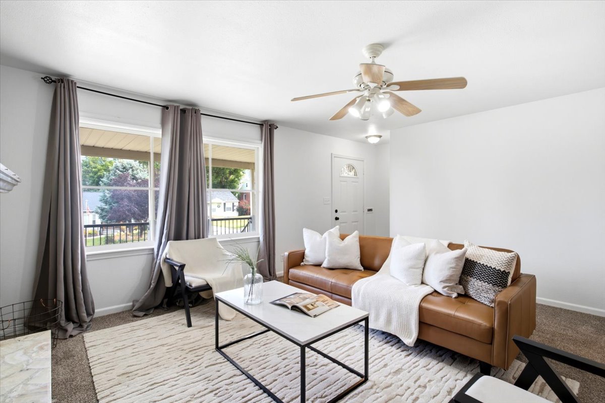 Living area featuring a ceiling fan with light fixture, carpet flooring, a white paneled entry door, and a pair of large windows overlooking the exterior porch