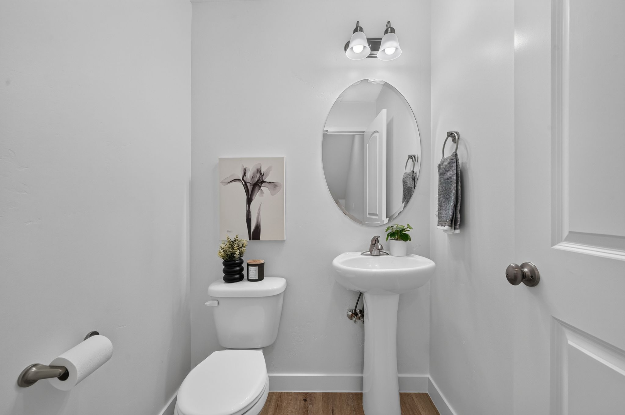 Pedestal sink with an oval mirror and brushed nickel hardware, complemented by wood-finish flooring and a two-light vanity fixture