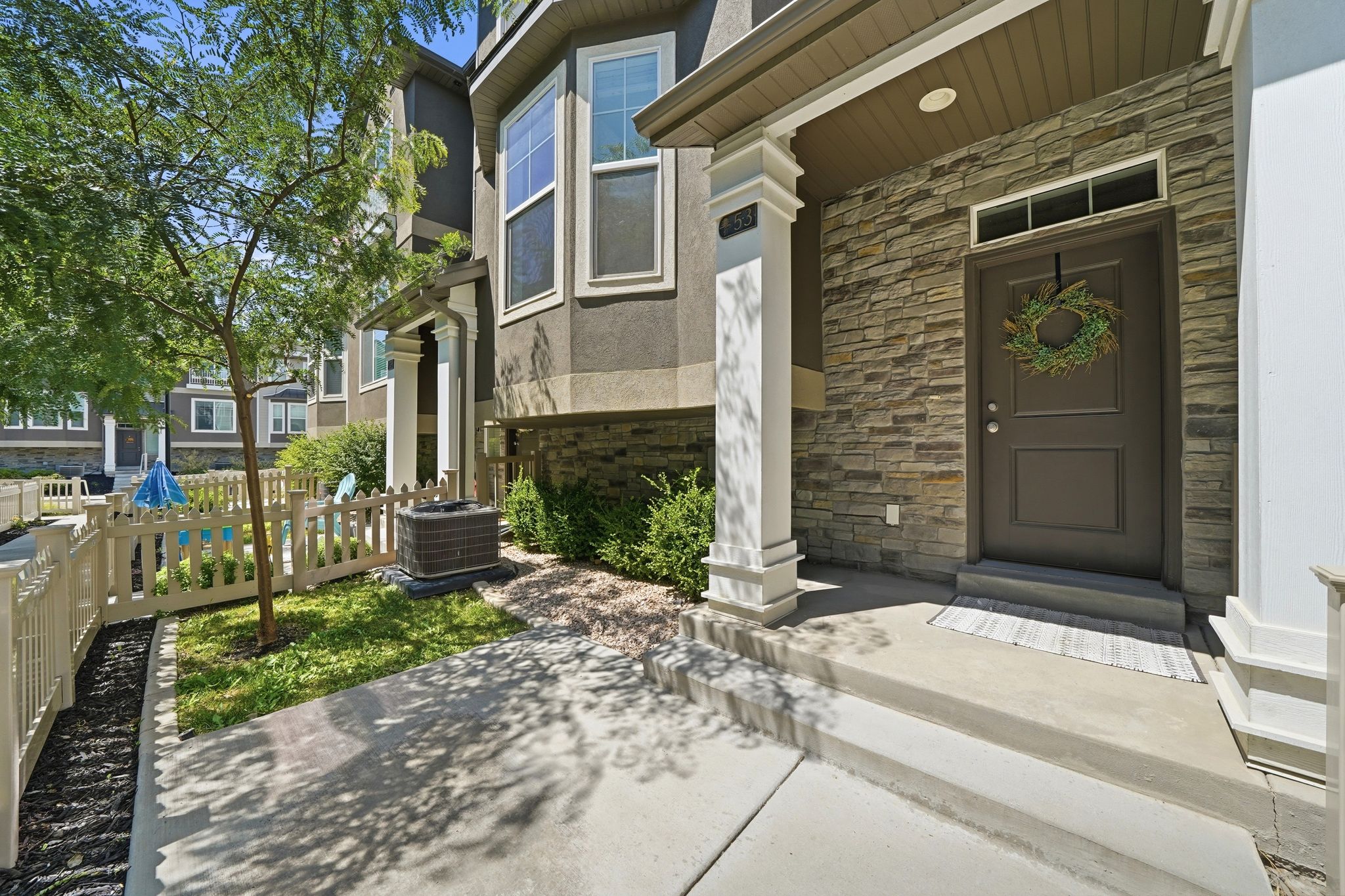 Front exterior featuring a stone-clad entry, dark gray door with transom window, concrete porch with decorative pillars, and a picket-fenced yard with mature landscaping
