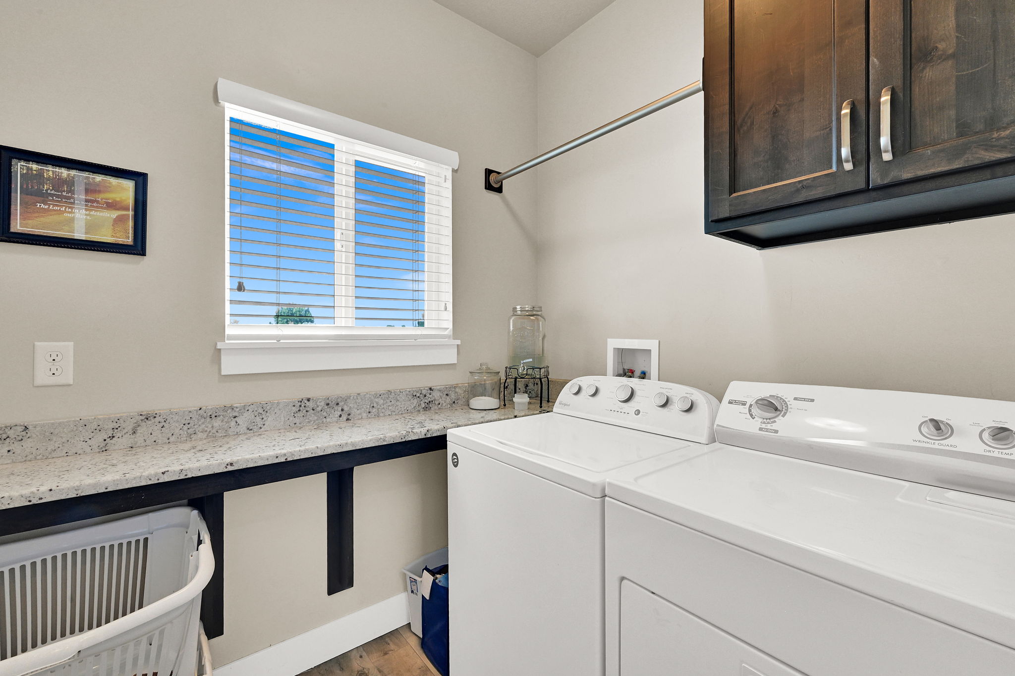 Dedicated laundry space featuring a window with blinds, granite-look countertops, dark wood-finish cabinetry, a chrome hanging rod, and wood-finish flooring