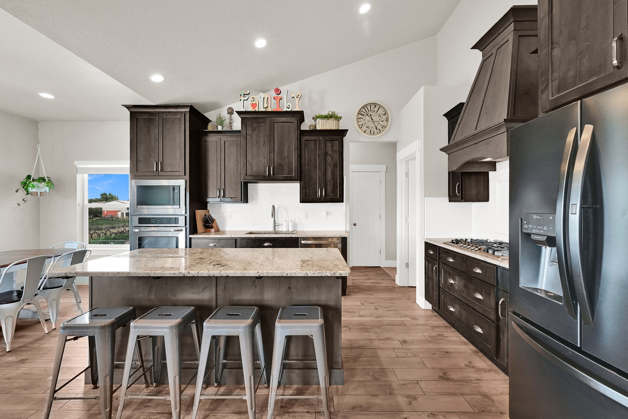 Open-concept kitchen featuring an oversized island with countertop seating, dark wood cabinetry, stainless steel appliances, and wood-finish flooring