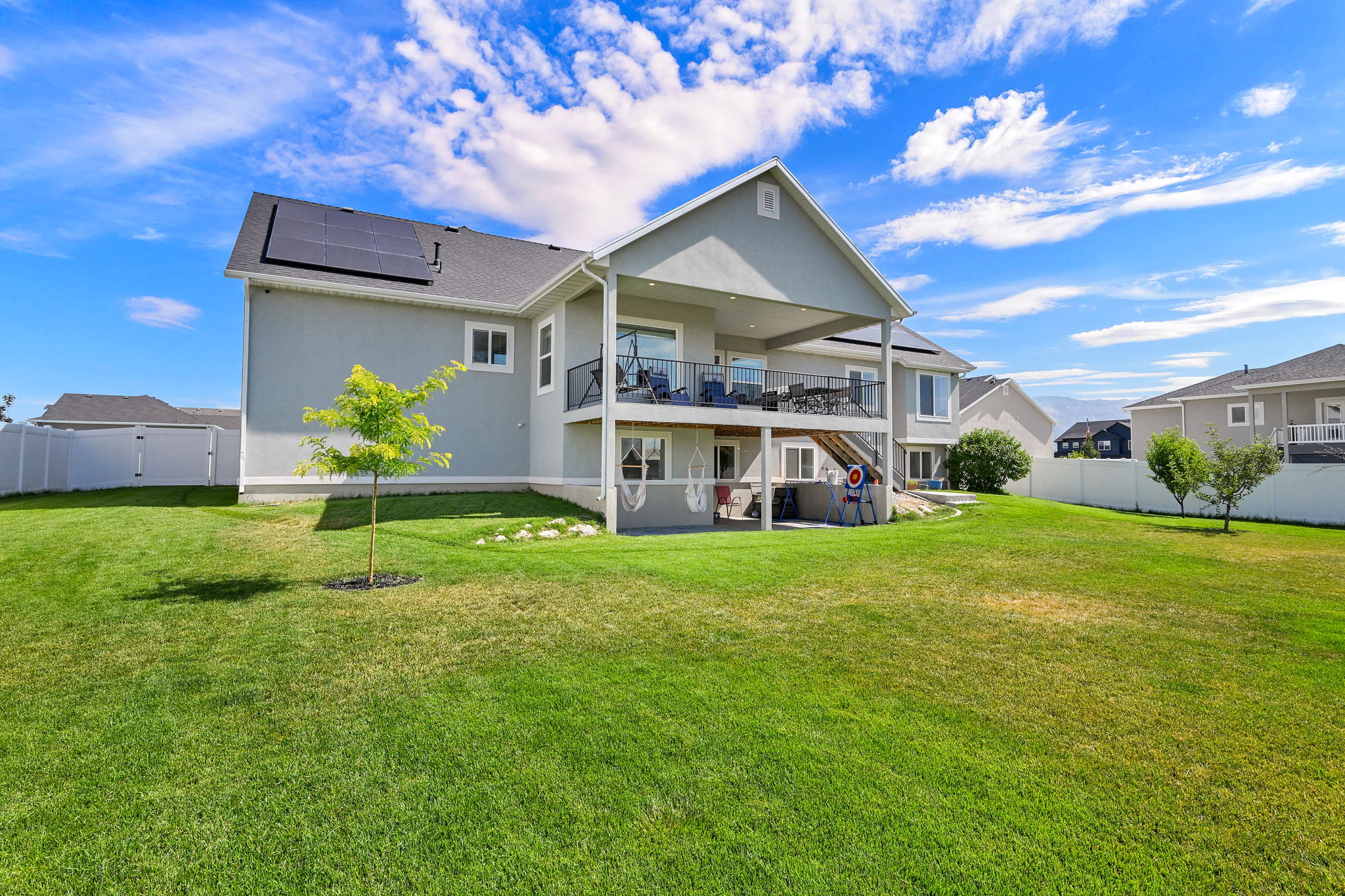 Two-story stucco residence featuring solar panels, a covered upper-level deck with black railings, and a covered lower-level patio