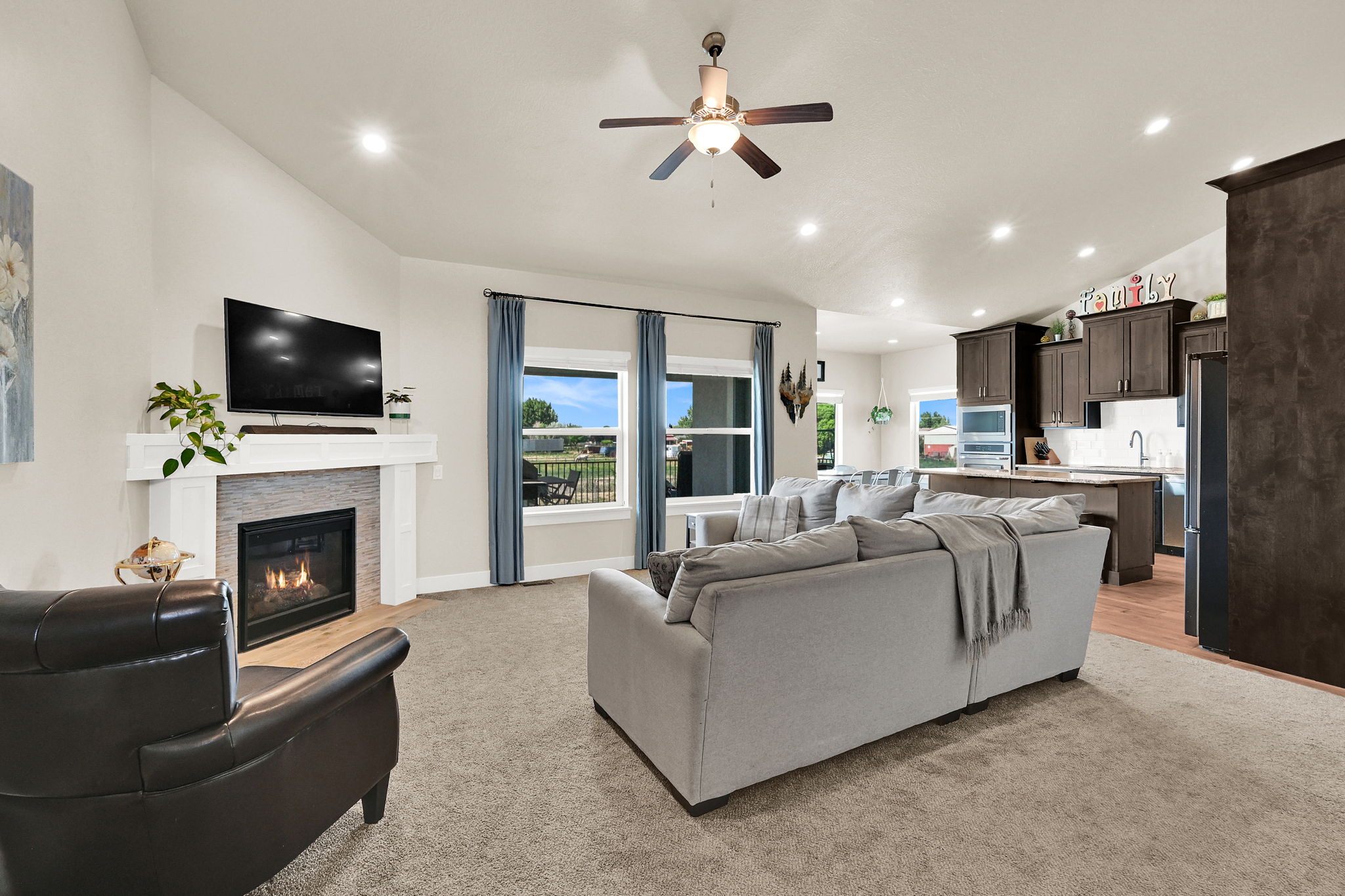 Open-concept living area featuring a gas fireplace with a stone-finish surround and white mantel, a ceiling fan with integrated lighting, recessed lighting, and large windows with blue drapery