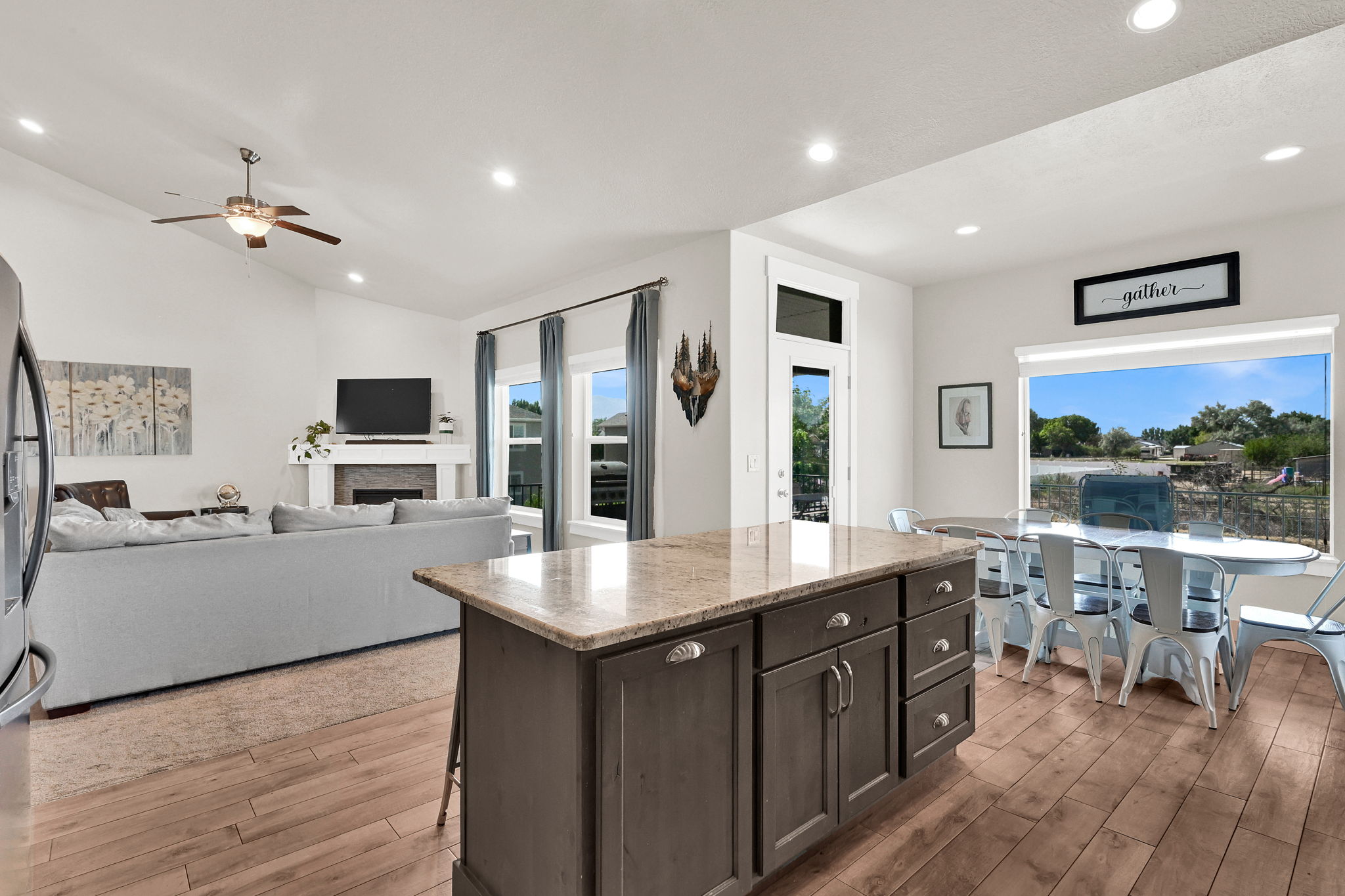 Open-concept living space featuring wood-finish flooring, a kitchen island with granite countertop and storage, a dining area with expansive window, a living room with a fireplace, and a vaulted ceiling with a ceiling fan