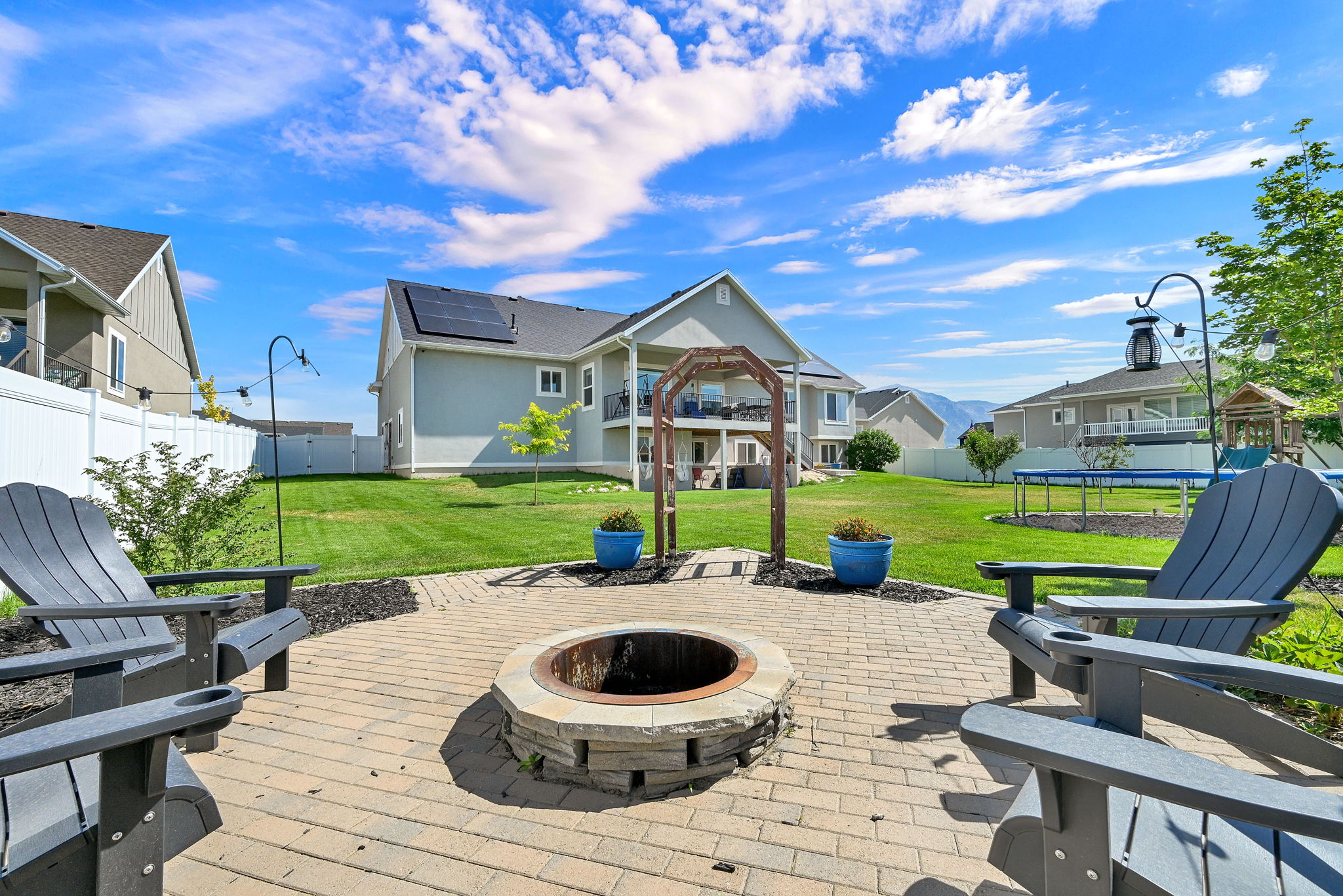 Expansive backyard featuring a paver patio with a built-in fire pit, manicured lawn, white privacy fencing, and a two-story residence with solar panels