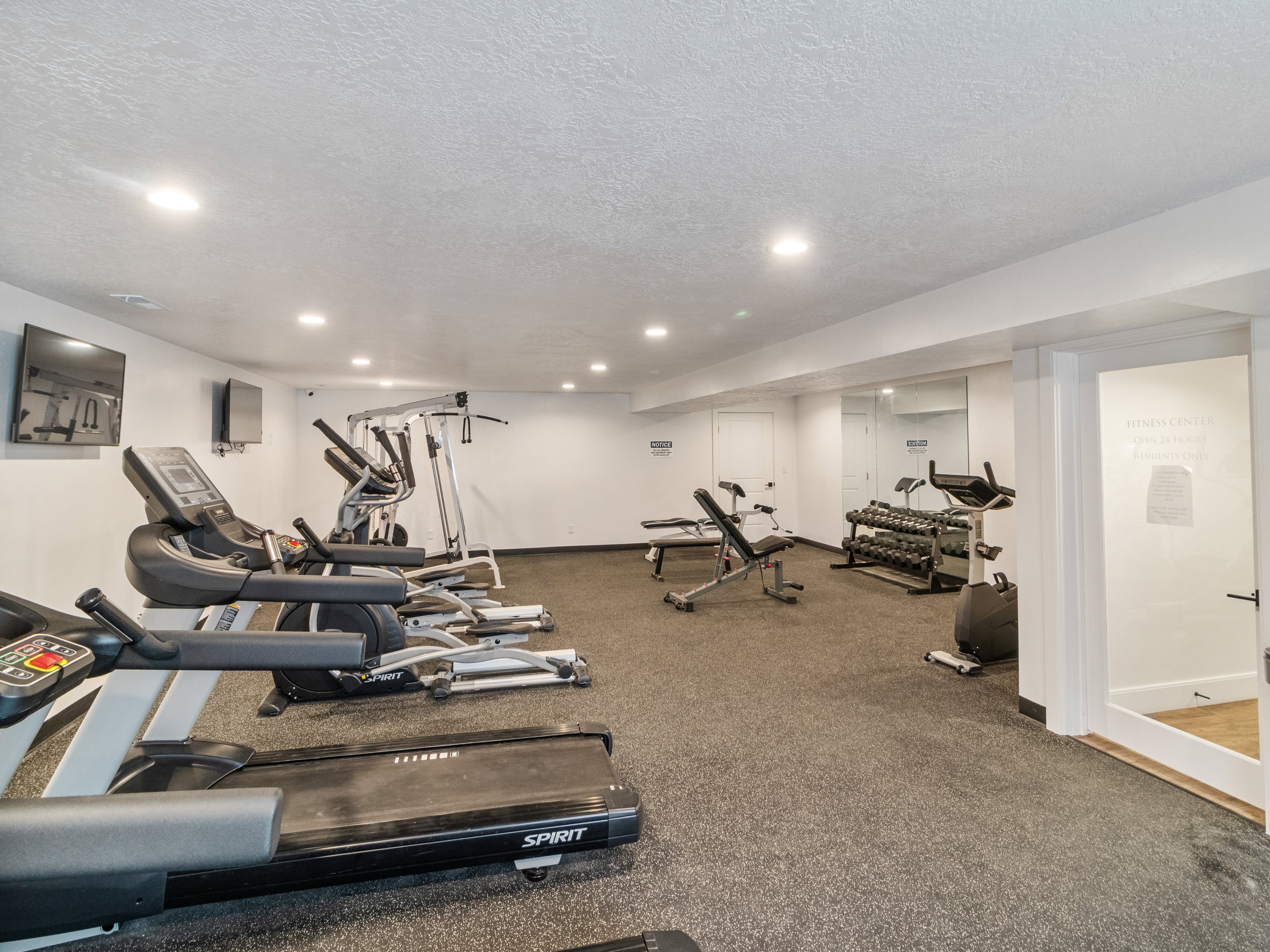 Clubhouse. Spacious fitness area featuring wall-mounted televisions, mirrored accent wall, recessed lighting, and resilient flooring