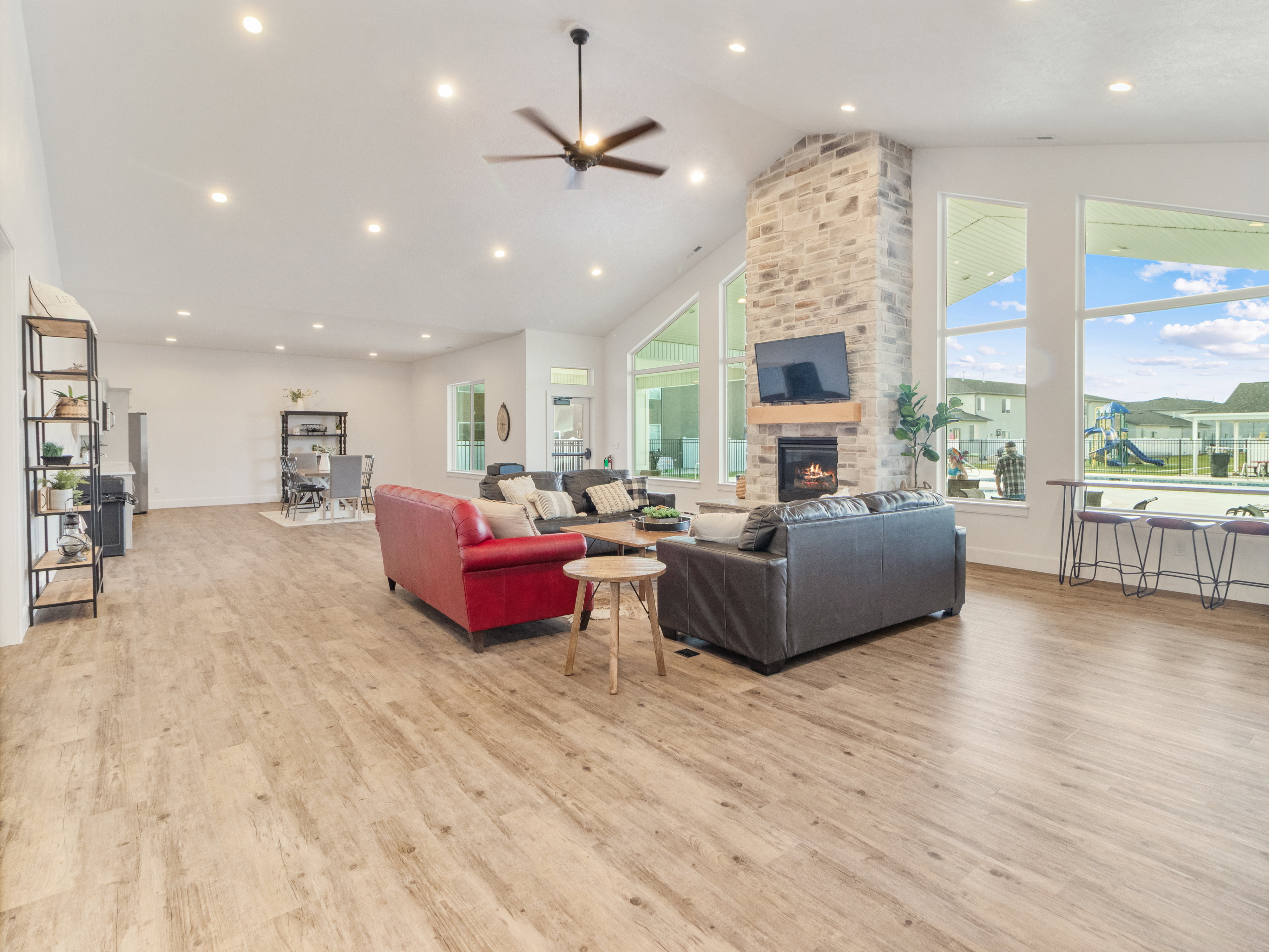 Clubhouse. Spacious open-concept living area featuring a stone-clad fireplace with a wood mantel, high vaulted ceilings, extensive window walls, recessed lighting, and wood-finish flooring