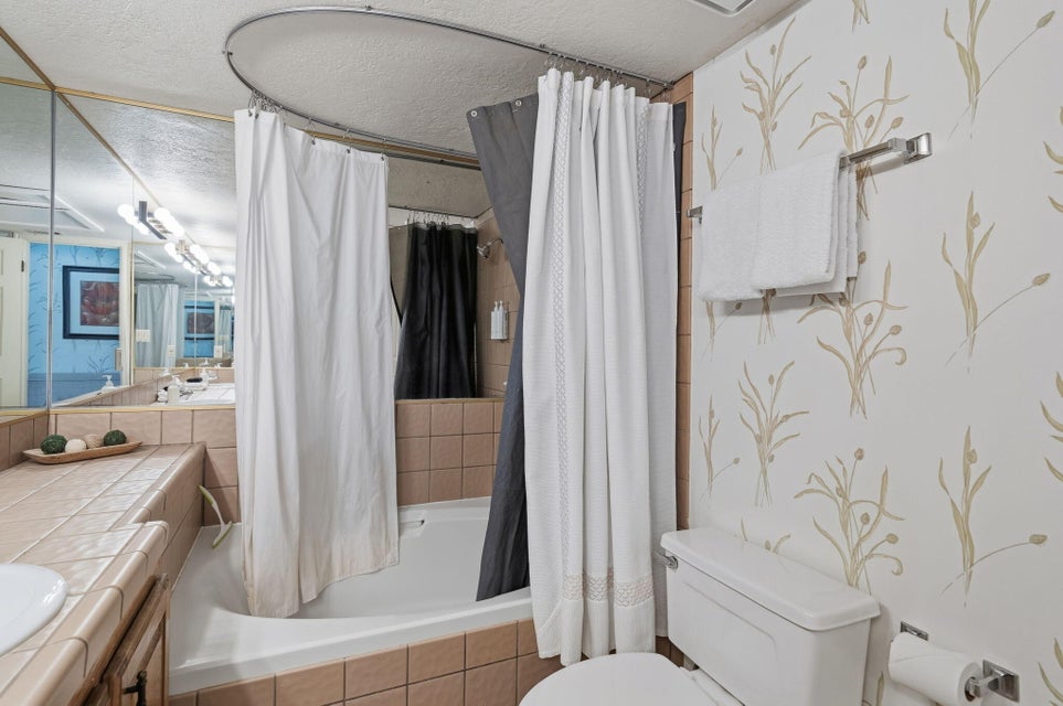 Bathroom featuring a built-in tub, extensive vanity countertop with tile finish, full-width mirror, and botanical-patterned wall covering