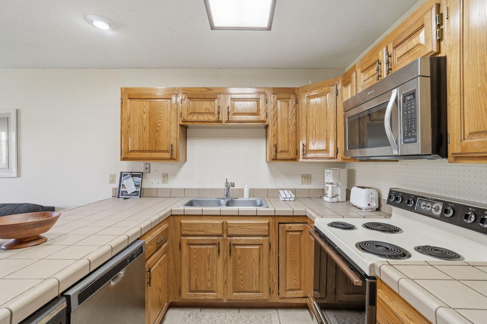 U-shaped kitchen featuring wood cabinetry, tile countertops, a double-basin stainless steel sink, a stainless steel microwave, and a white electric range