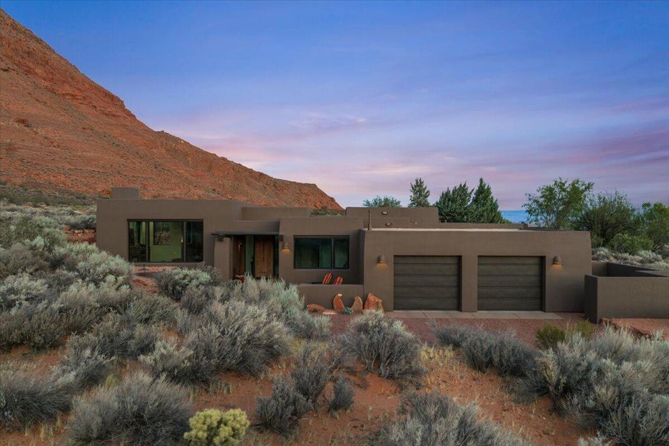 Southwestern-style residence featuring a stucco exterior, integrated two-car garage with dark wood-finish doors, prominent rock formations, and arid landscaping
