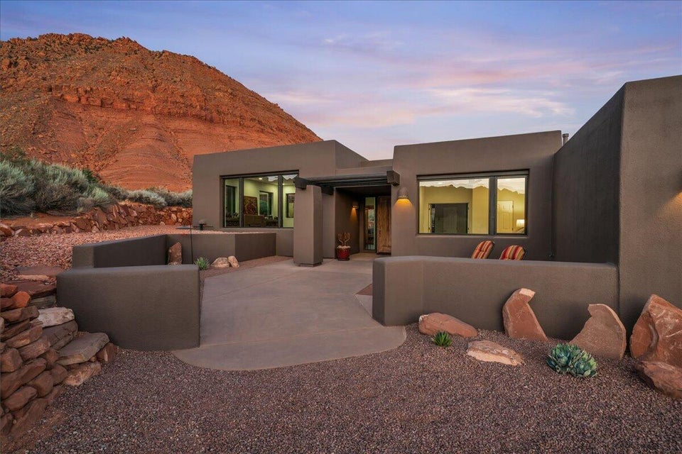 Desert contemporary architecture with a stucco exterior, expansive windows, a concrete patio, integrated outdoor lighting, and natural rock landscaping