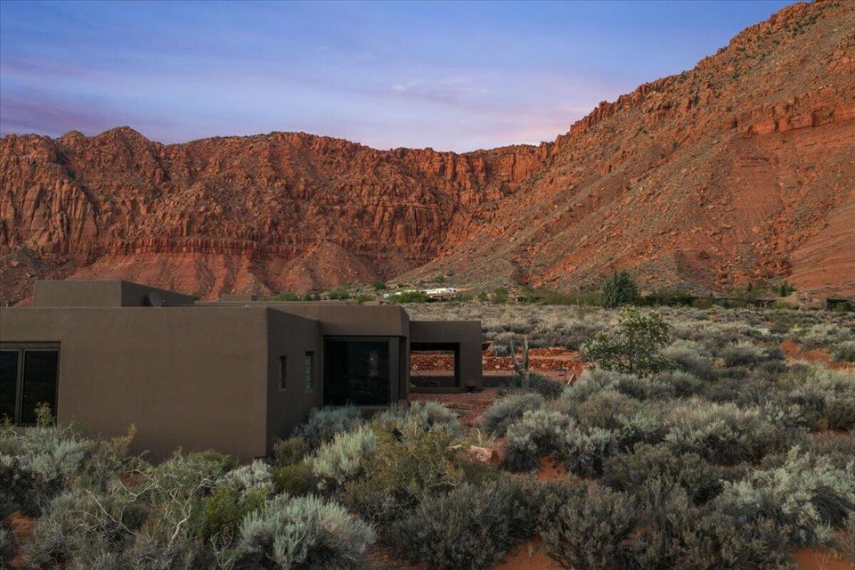 Modern-style exterior with a flat roofline, stucco finish, and dark-framed windows, surrounded by natural desert landscaping and dramatic red rock formations