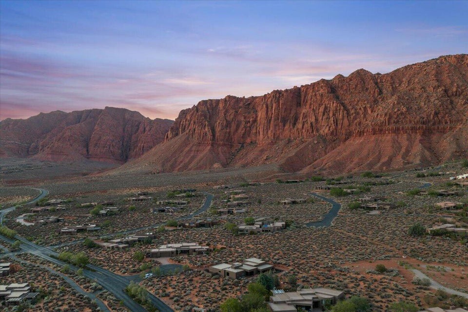 Expansive desert landscape featuring a residential community with contemporary low-profile homes, winding roadways, and mature desert vegetation set against dramatic red rock formations