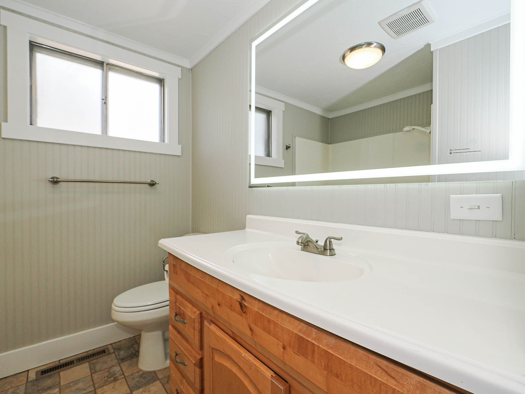 Bathroom featuring a wood-finish vanity with a white integrated sink, an illuminated mirror, and a window for natural light