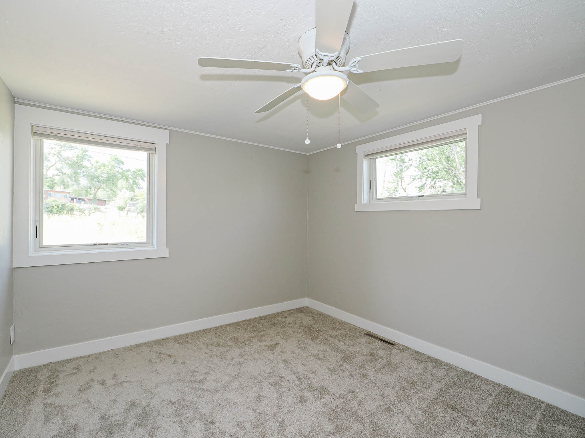 Neutral-toned room featuring a ceiling fan with integrated lighting, two windows with white trim, light gray walls, and textured gray carpeting