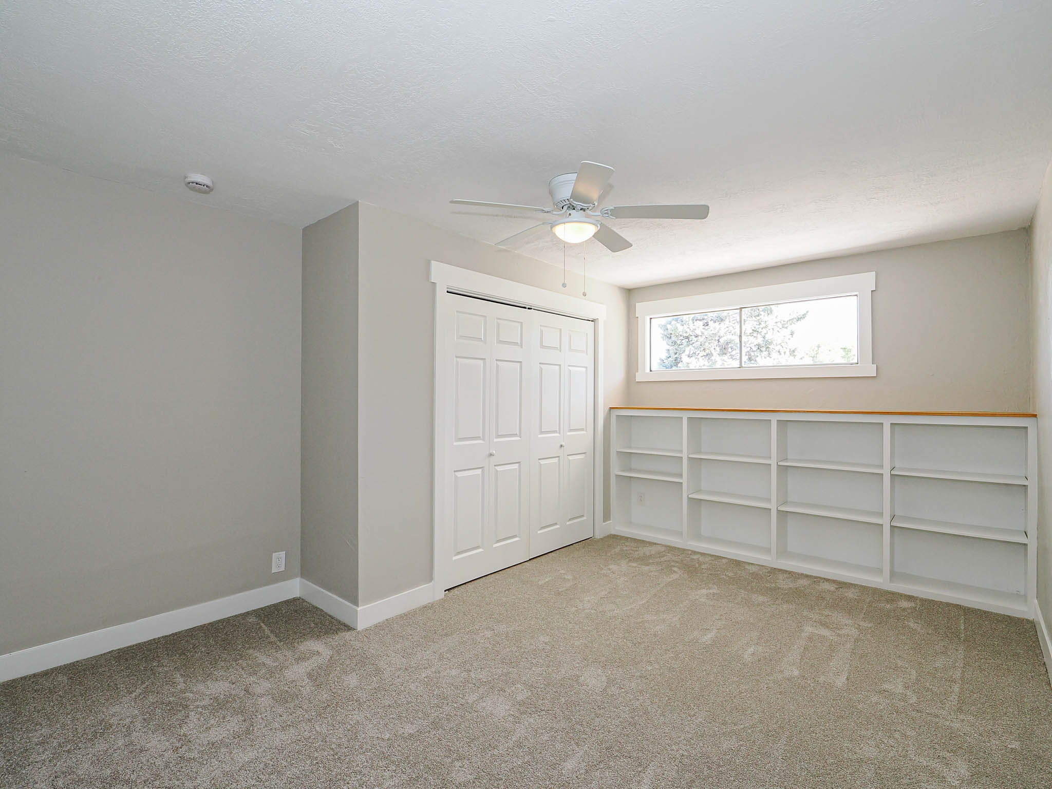 Spacious room featuring neutral carpeting, a ceiling fan, a built-in closet with bifold doors, and extensive built-in shelving with a light wood-finish top
