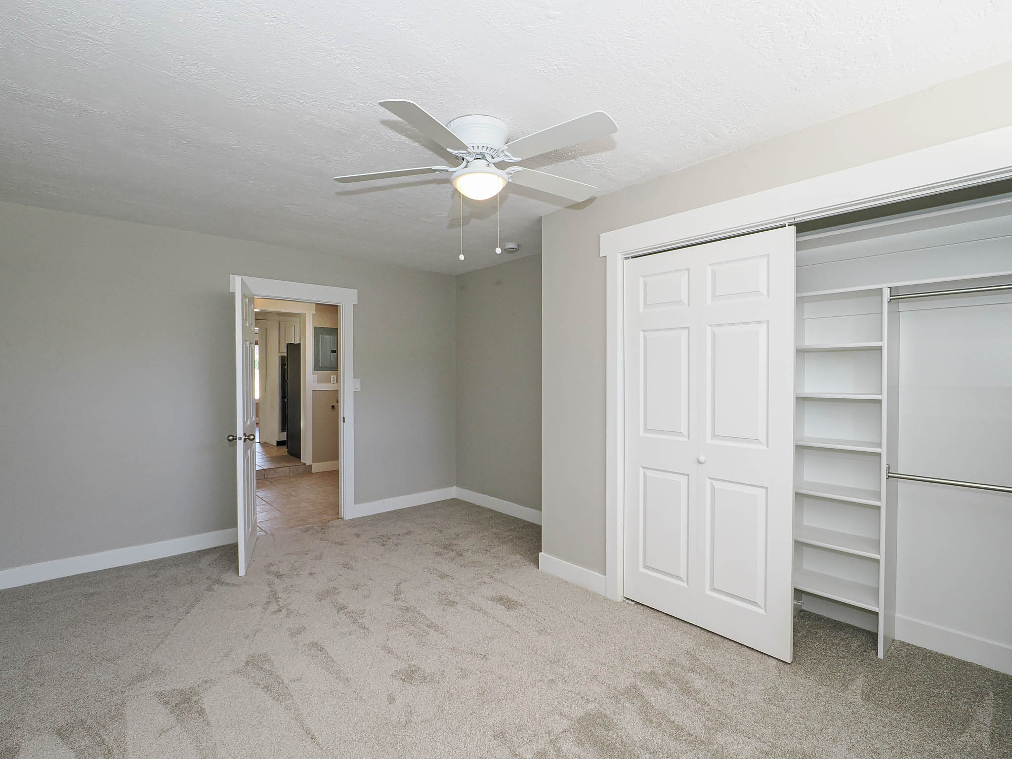 Neutral-toned carpeted room featuring a ceiling fan with integrated lighting, a sliding closet with built-in shelving, and a white paneled door