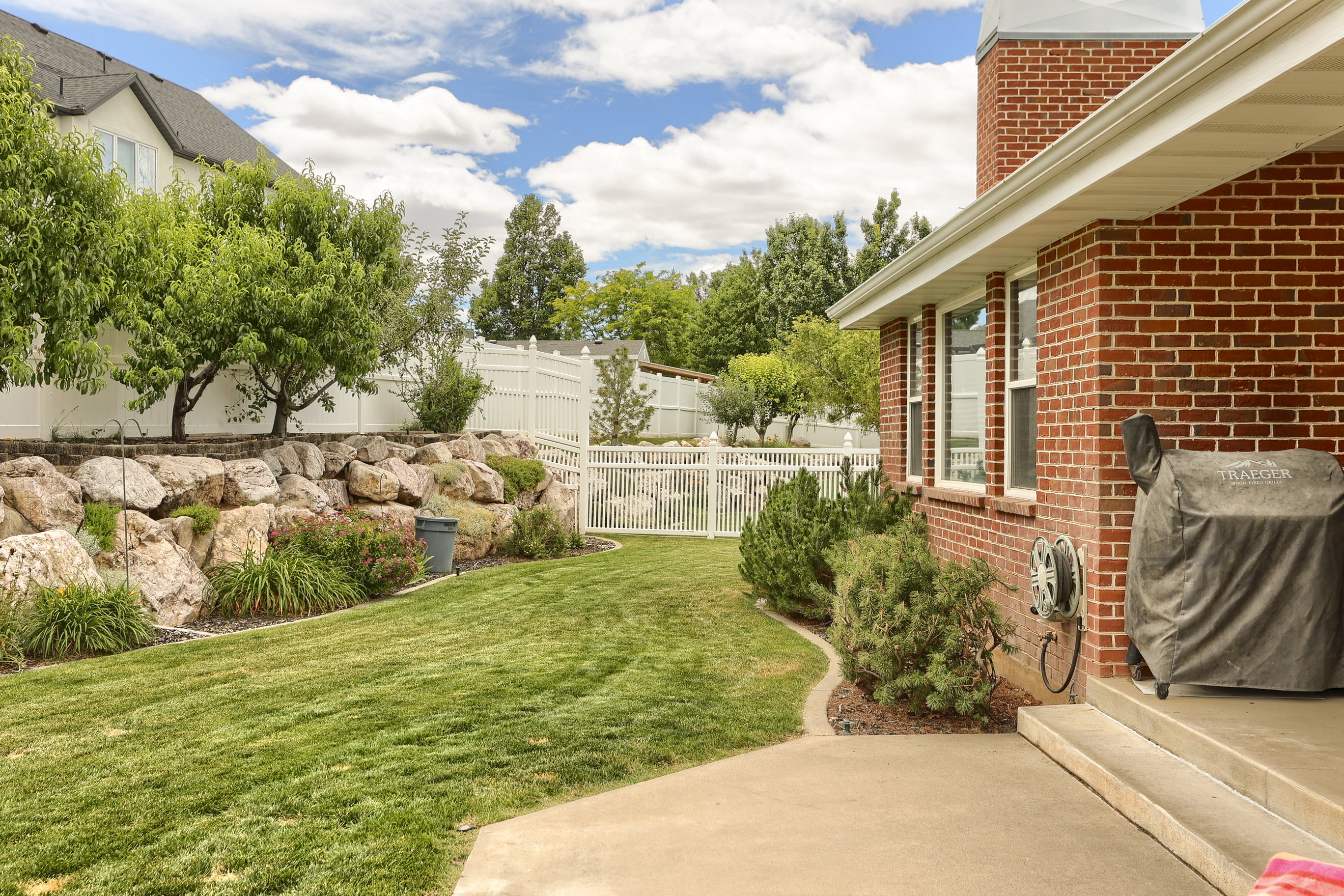 Expansive backyard featuring a manicured lawn, a rock retaining wall, mature trees, and a white privacy fence