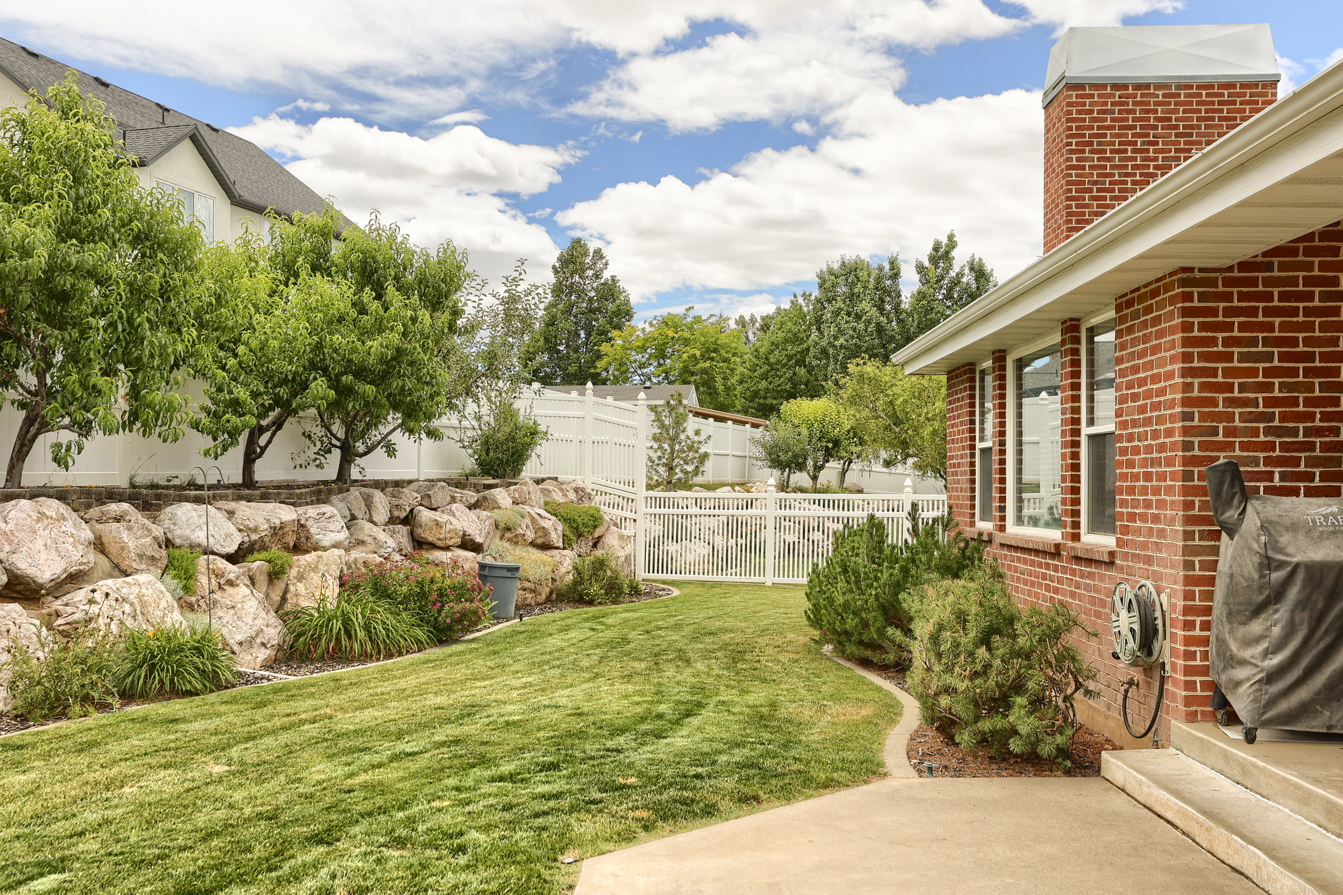 Expansive backyard featuring a lush lawn, a concrete patio, a red brick exterior, and a rock retaining wall with mature trees and shrubbery