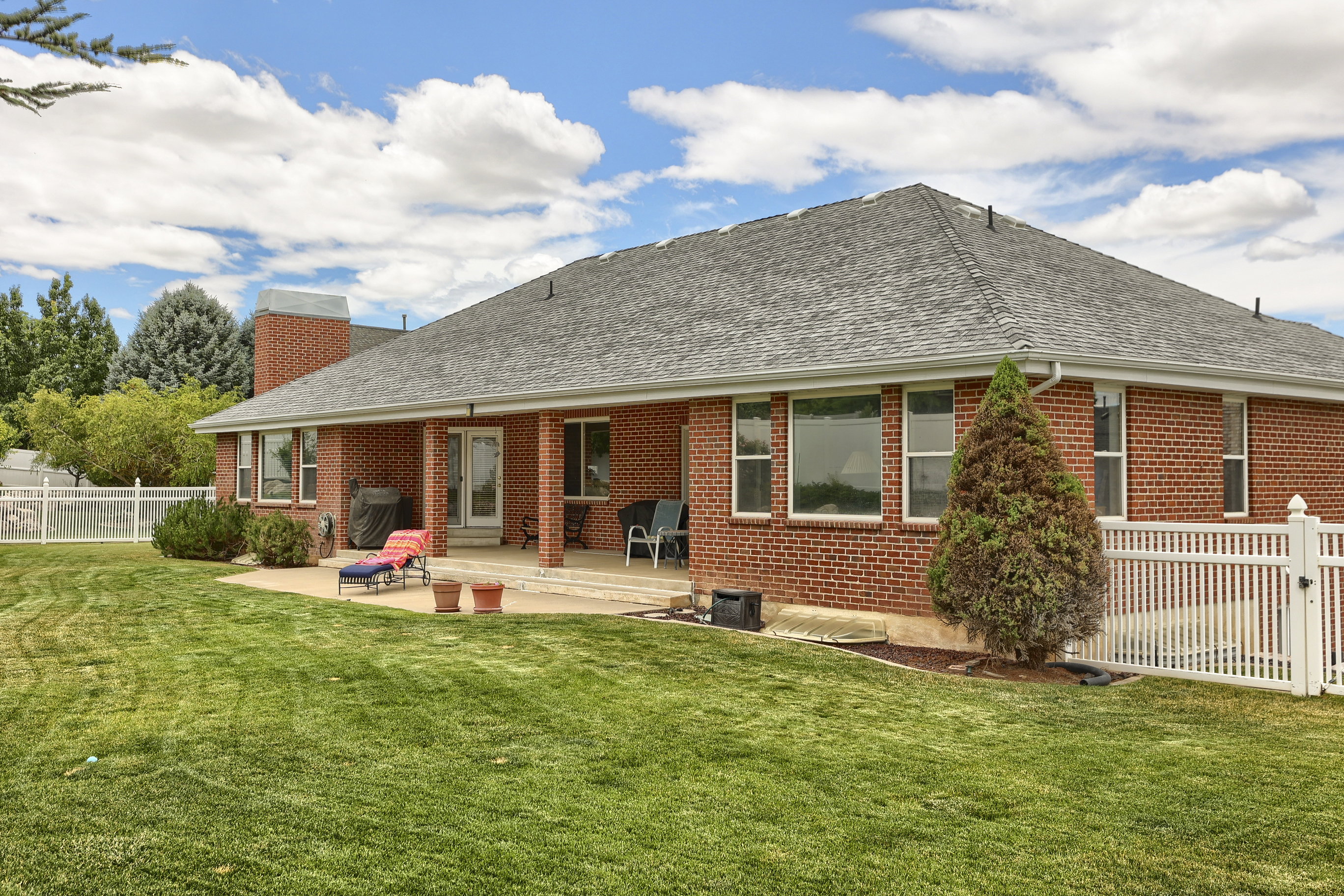 Expansive green lawn bordering a brick exterior with a covered patio area, featuring multiple windows and a white picket fence