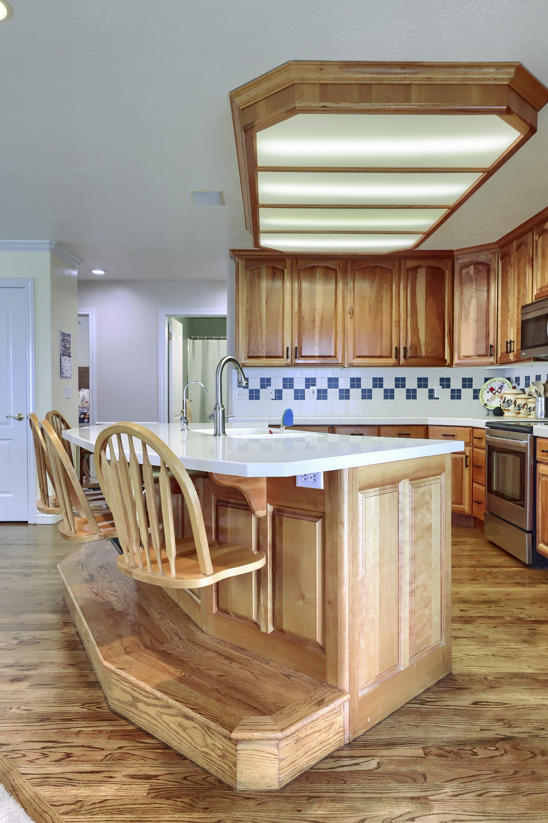 Kitchen featuring a large central island with integrated seating, light wood-finish cabinetry, a patterned tile backsplash, and a large tray ceiling light fixture