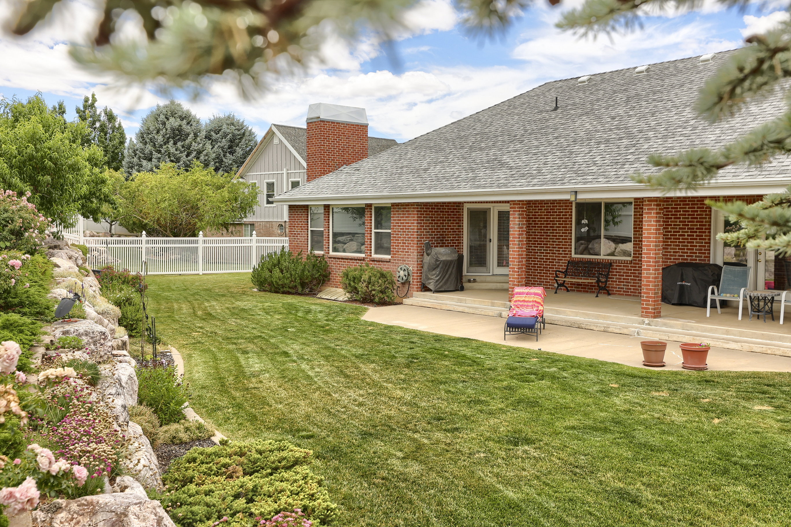 Expansive backyard featuring a manicured lawn, established rock landscaping with flowering shrubs, and a rear patio