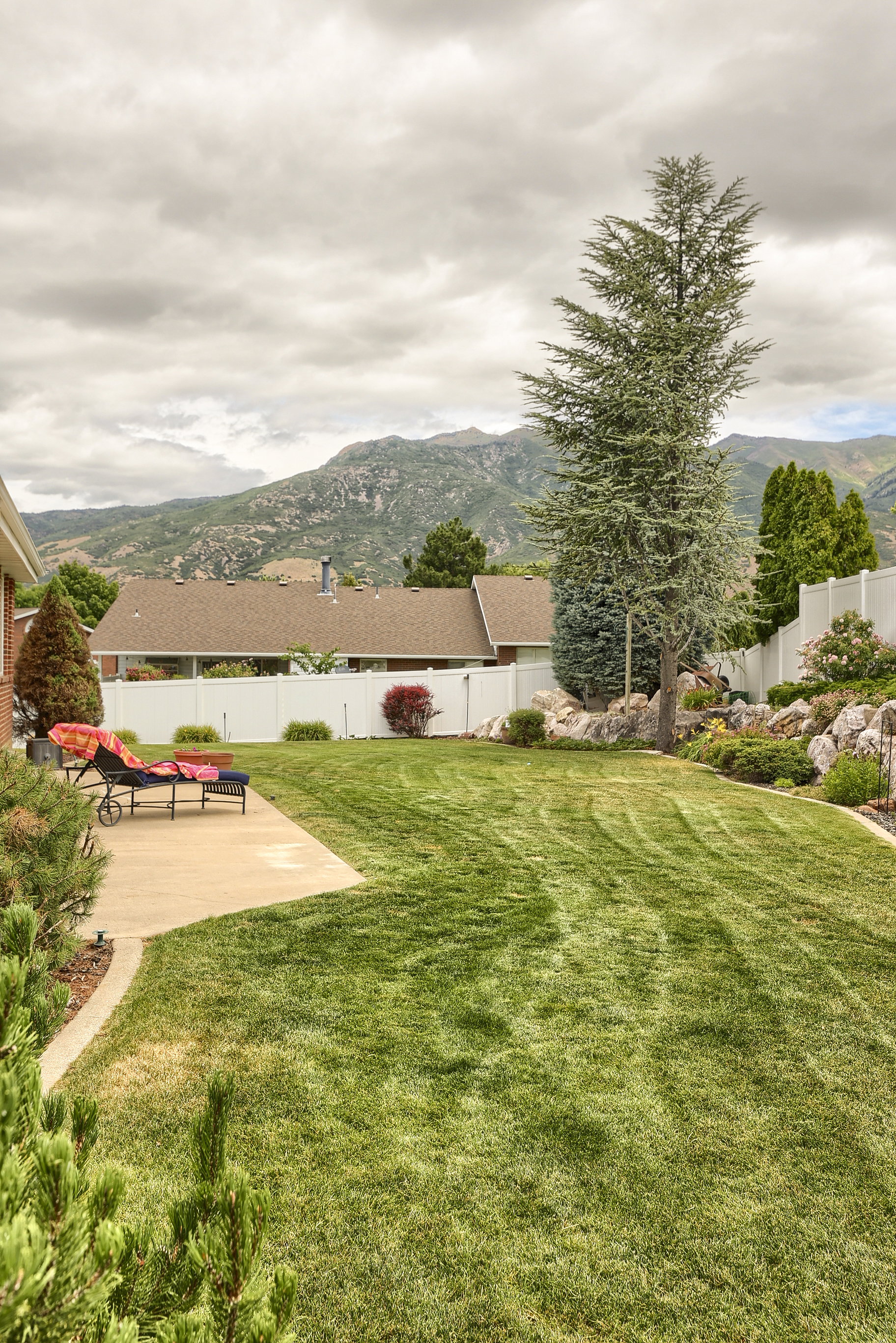 Expansive backyard featuring a lush lawn, concrete patio, mature evergreen trees, and a white privacy fence