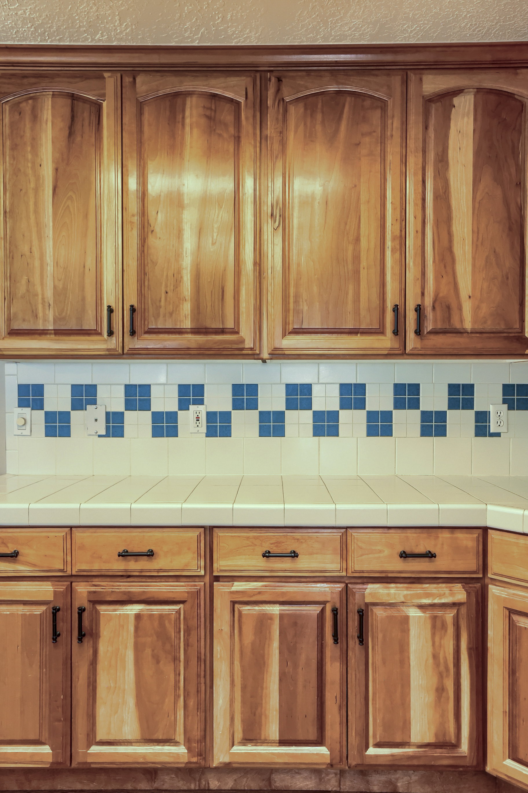 Natural wood cabinetry with arched top panels, a light-toned tiled countertop, and a white tiled backsplash featuring a blue accent border