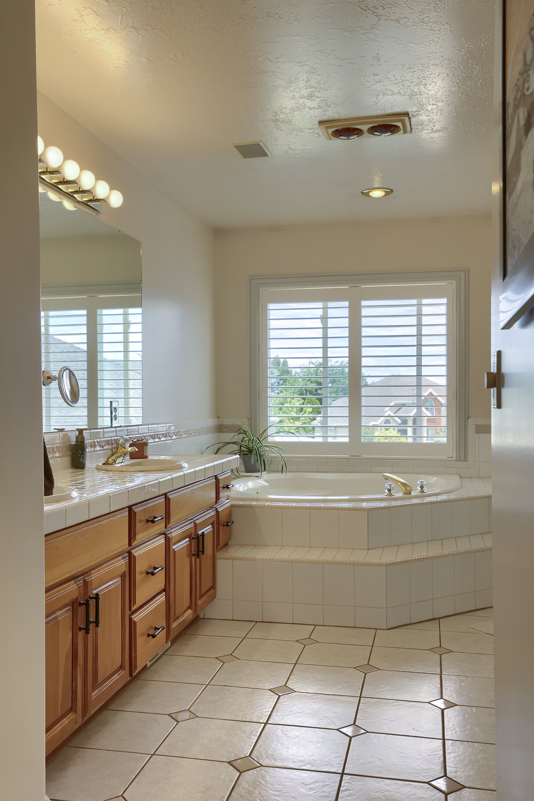 Corner soaking tub with a tiled surround, plantation shutters, and an adjacent vanity with wood cabinetry and a tile countertop
