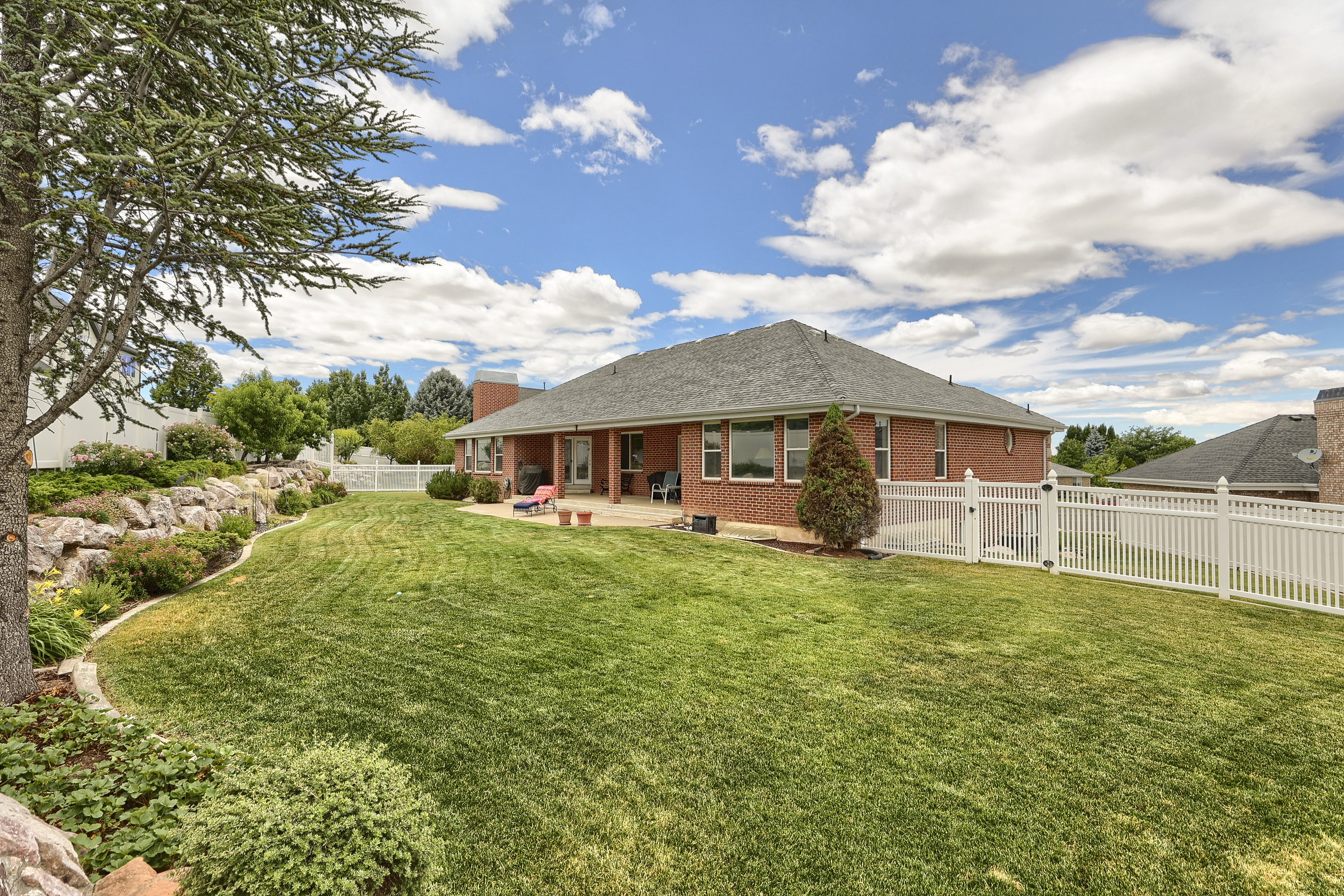 Expansive backyard featuring a manicured lawn, a white picket fence, a rock retaining wall with mature landscaping, a paved patio, and a brick exterior with multiple windows
