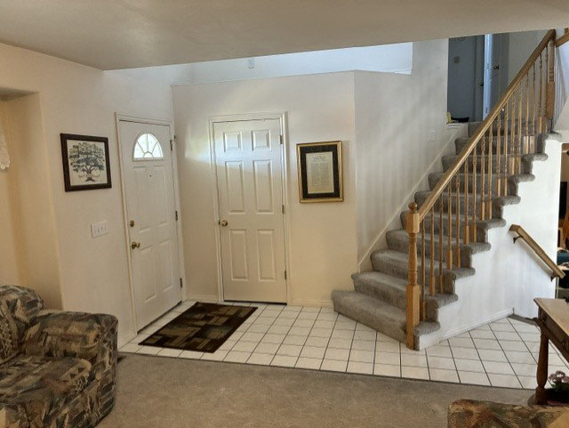 Entry foyer with neutral tile flooring, a carpeted staircase with wood banister, and two white paneled doors