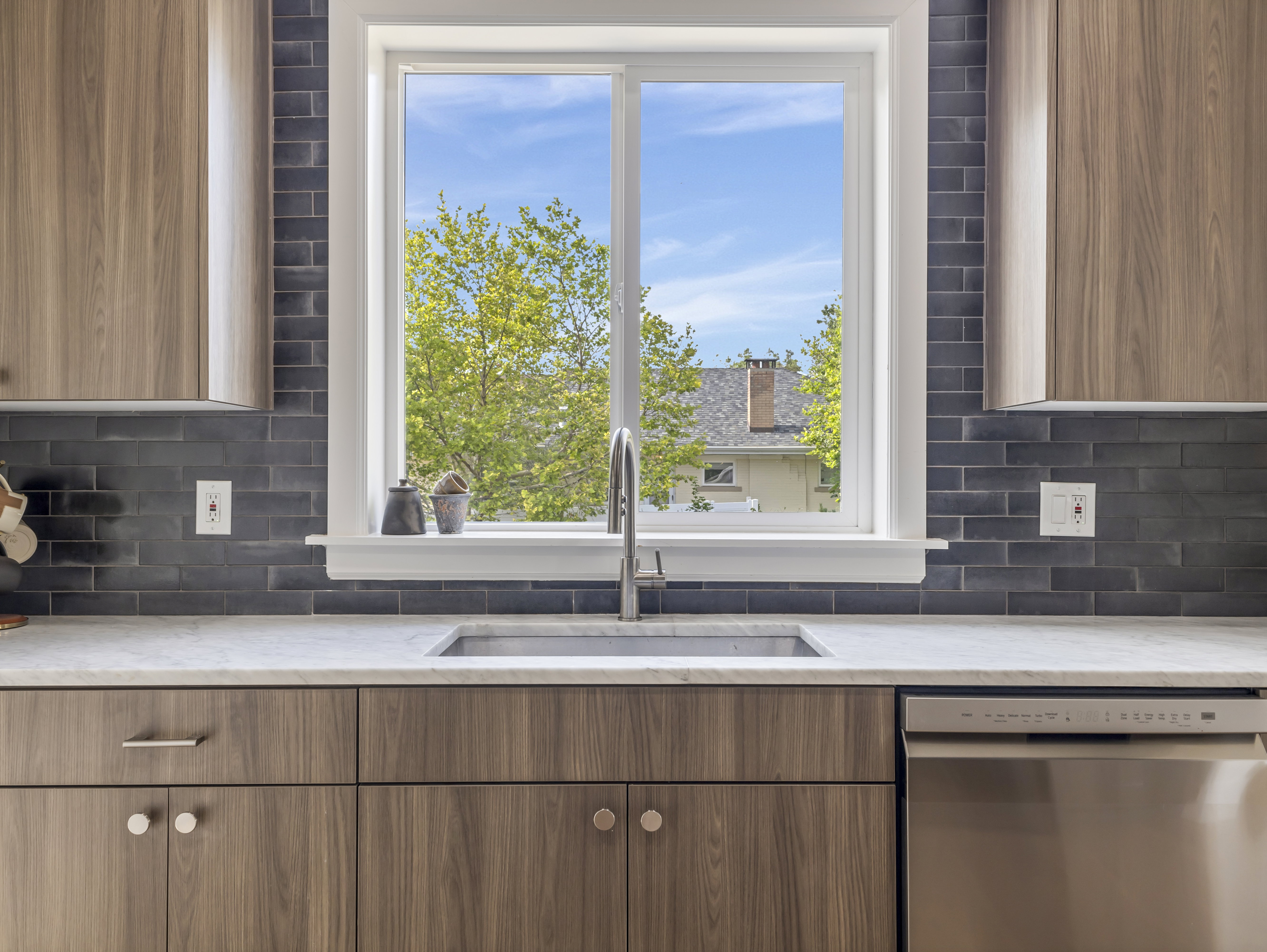 Kitchen featuring a stainless steel sink with a gooseneck faucet, light-colored countertop, dark subway tile backsplash, wood-finish cabinetry with various hardware, and a built-in stainless steel dishwasher