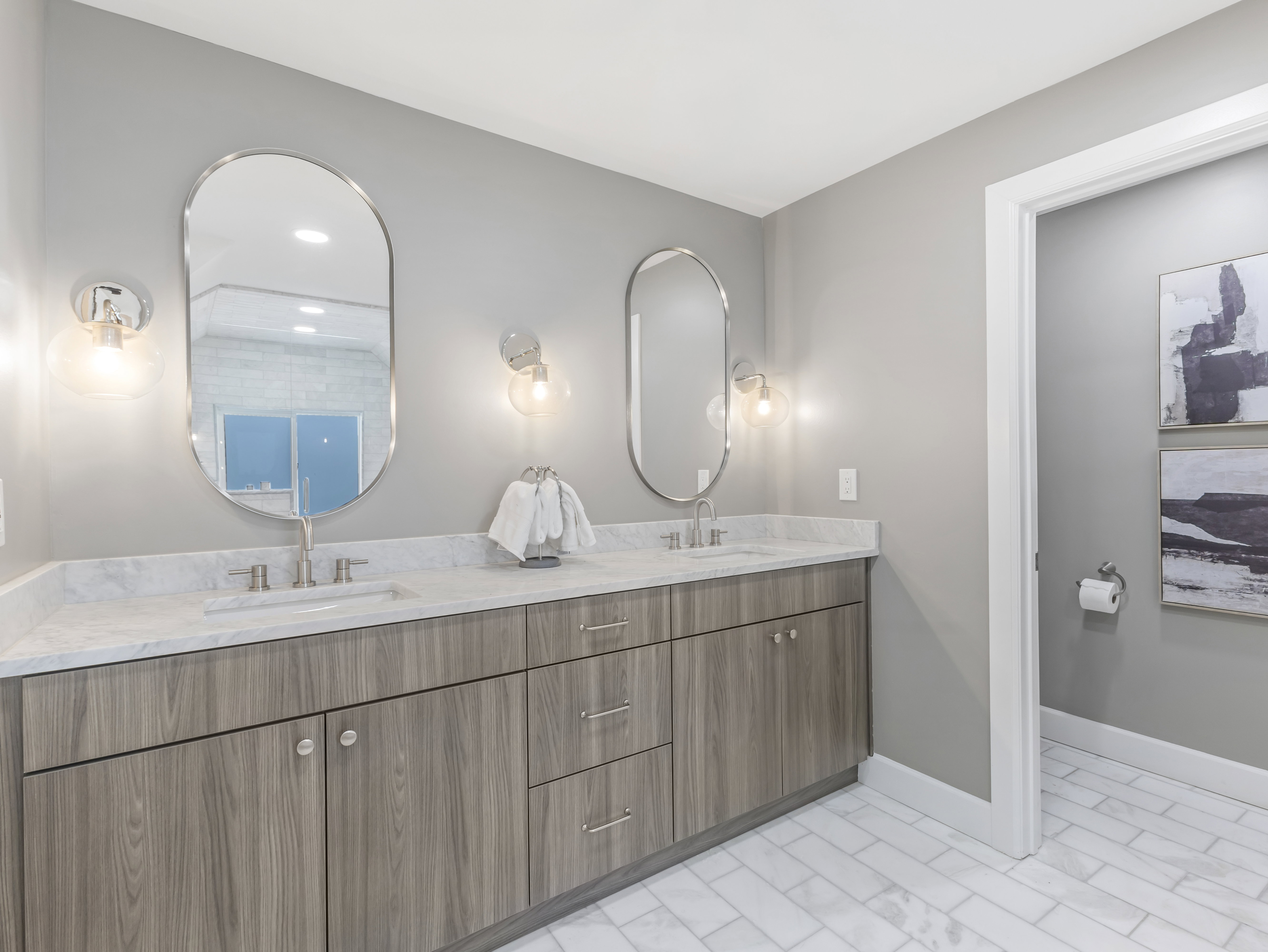 Double vanity featuring a light wood-finish cabinetry, white countertop, and dual oval mirrors with sconce lighting