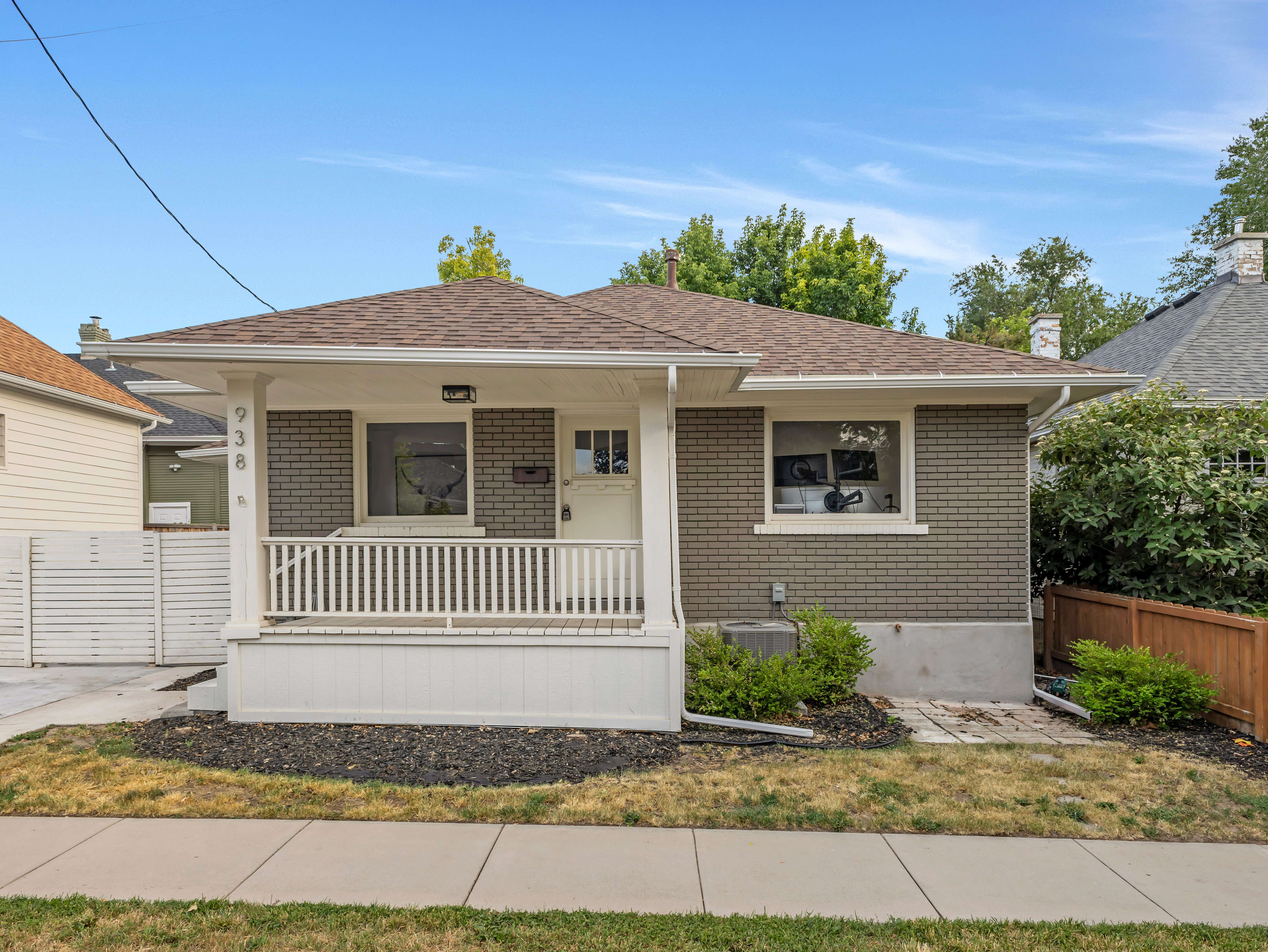Traditional bungalow exterior featuring gray brick siding, a covered front porch with white railing, a brown shingle roof, and established landscaping