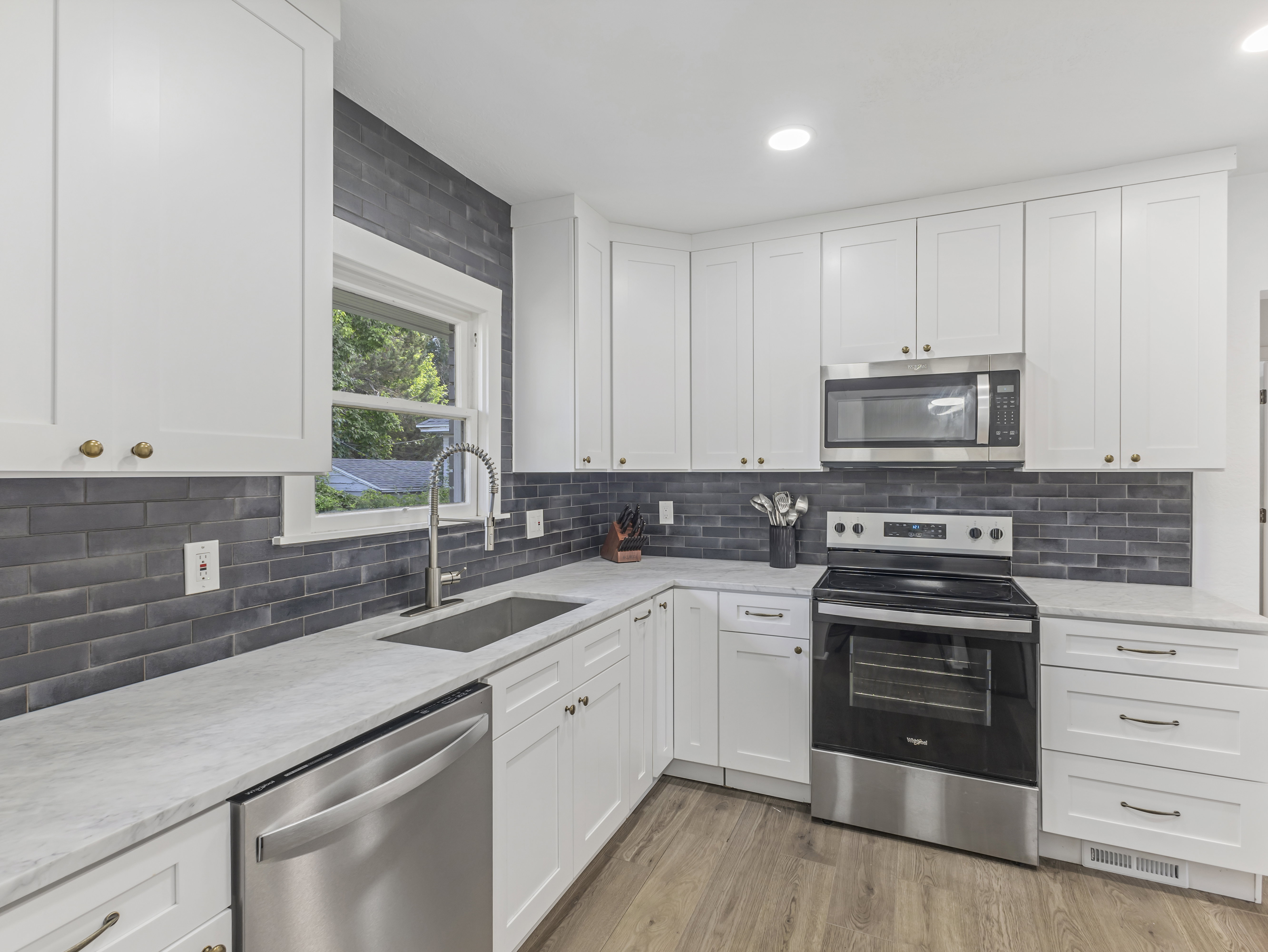 Modern kitchen showcasing white cabinetry with brass hardware, stainless steel appliances, a deep basin sink with a gooseneck faucet, light-toned countertops, and a dark tile backsplash
