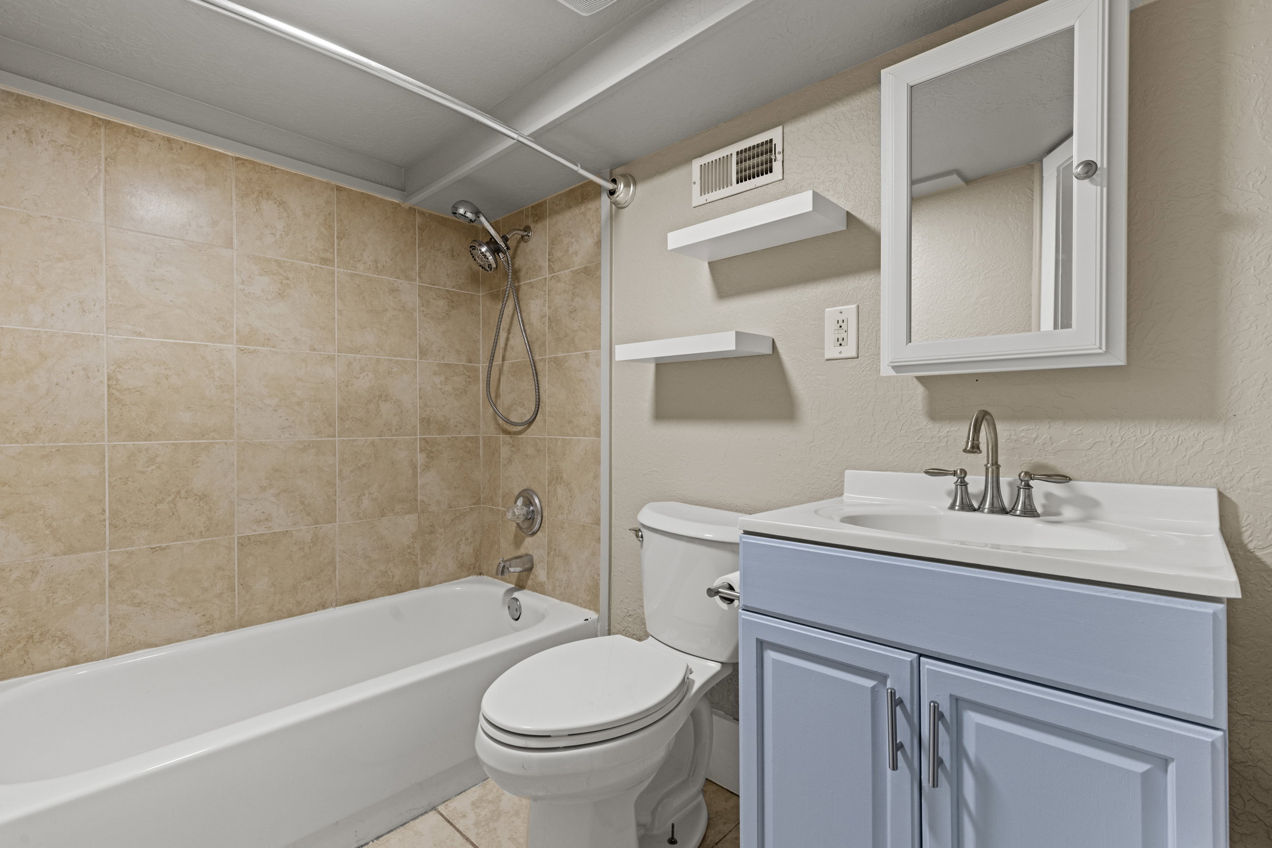 Bathroom featuring a white bathtub, beige tiled shower surround, toilet, light blue vanity with a white countertop and basin sink, and a mirrored medicine cabinet