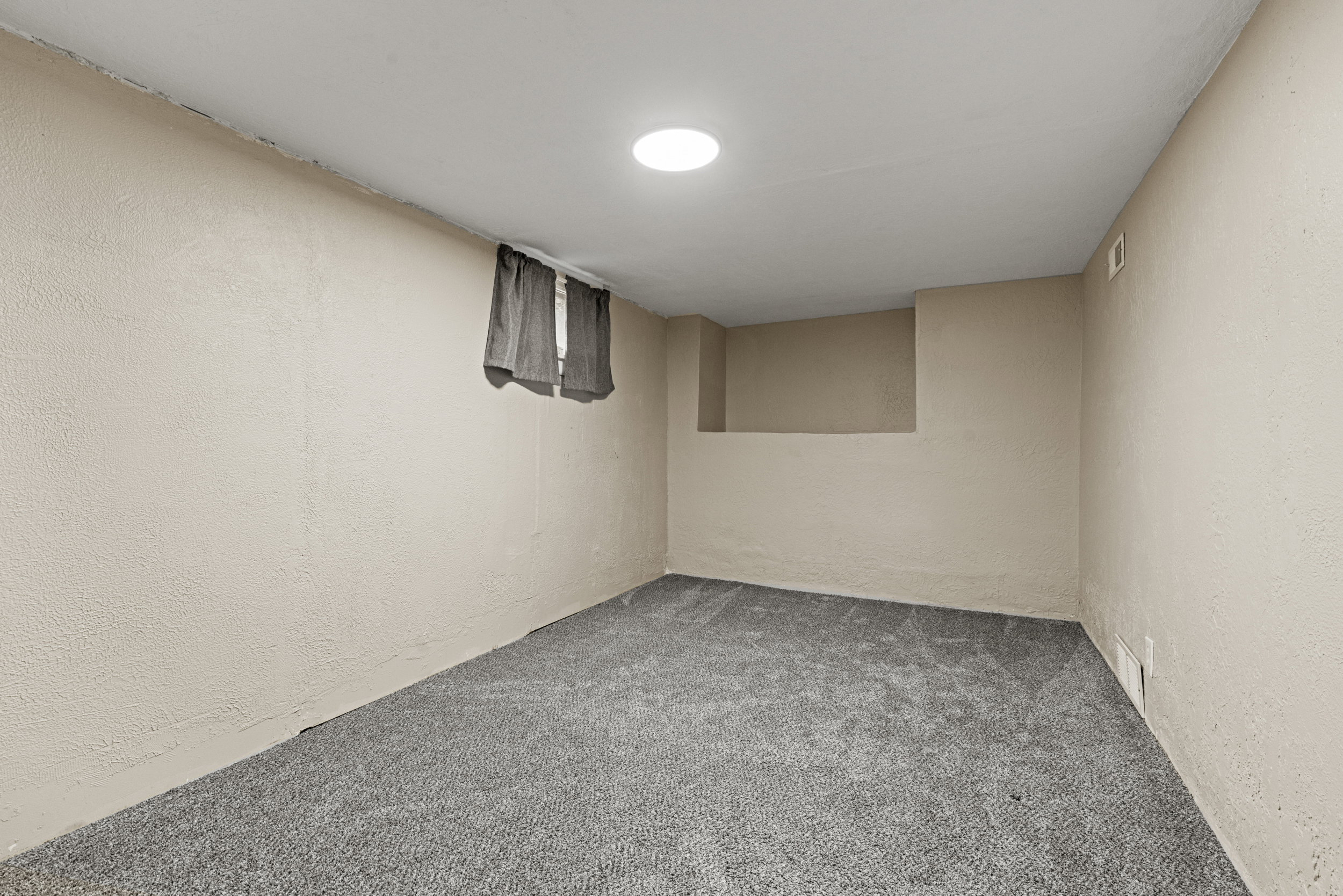 Carpeted room with a recessed circular light fixture, a small window with drapes, and a wall protrusion