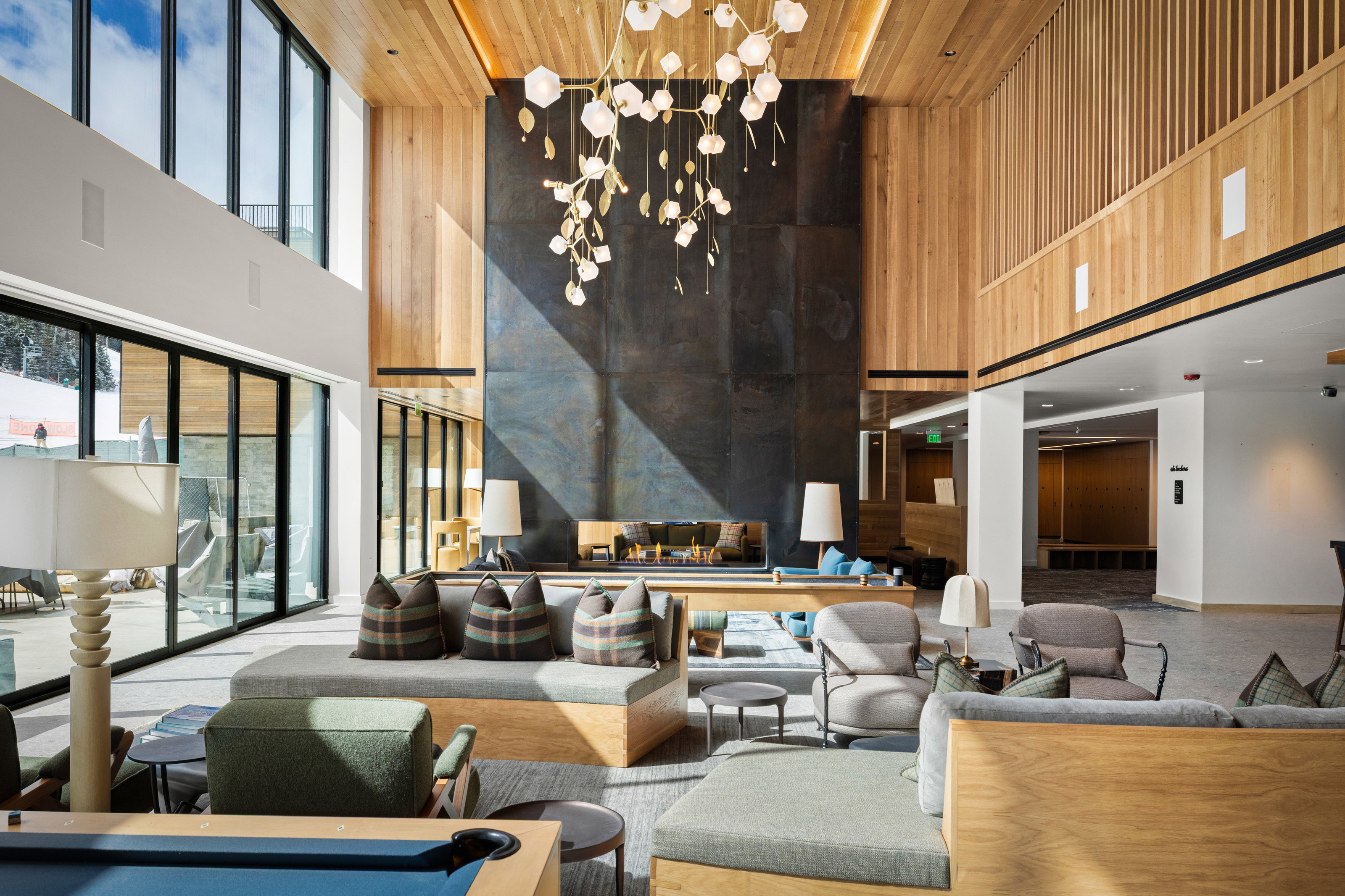 Expansive lounge featuring a dramatic two-story fireplace with dark paneling, wood-paneled walls and ceiling, a contemporary chandelier, a wall of sliding glass doors, and a built-in bench seating arrangement