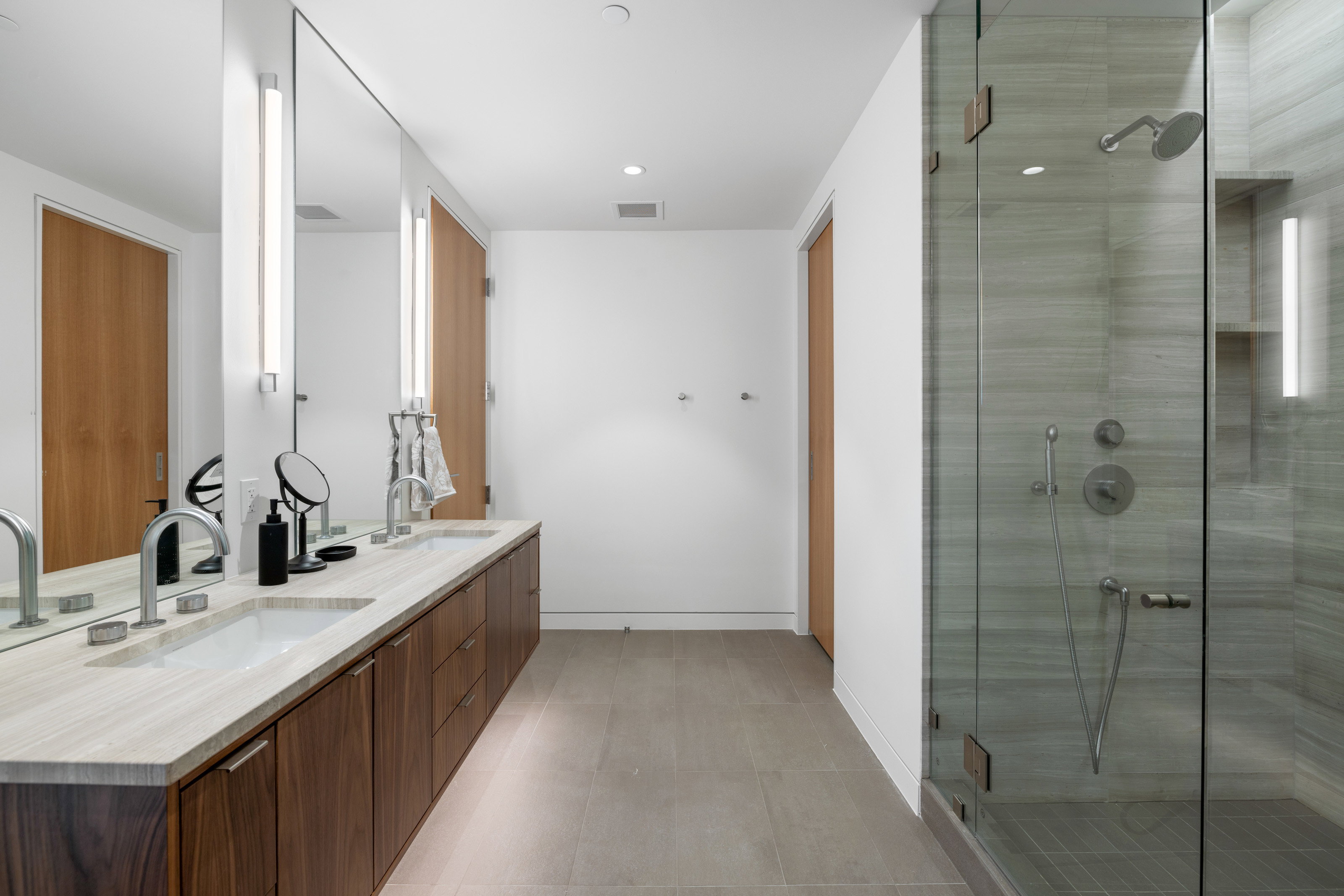 Spacious bathroom featuring a dual vanity with a light-toned countertop, wood-finish cabinetry, and a large frameless mirror